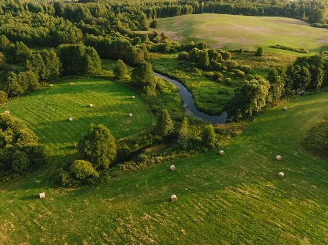 Aerial view of a green rural landscape with a winding creek, open fields with hay bales, and a dense forest.