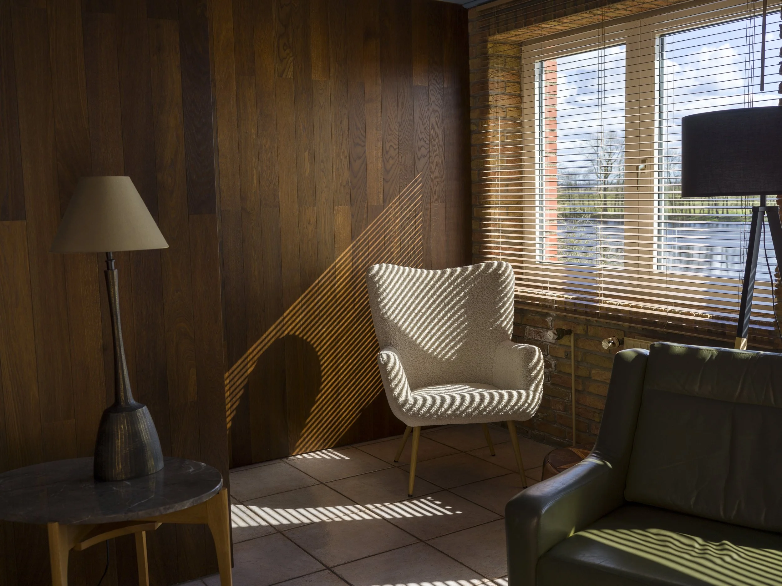 Sunlight streaming through window blinds onto a beige armchair and wooden wall in a cozy room.