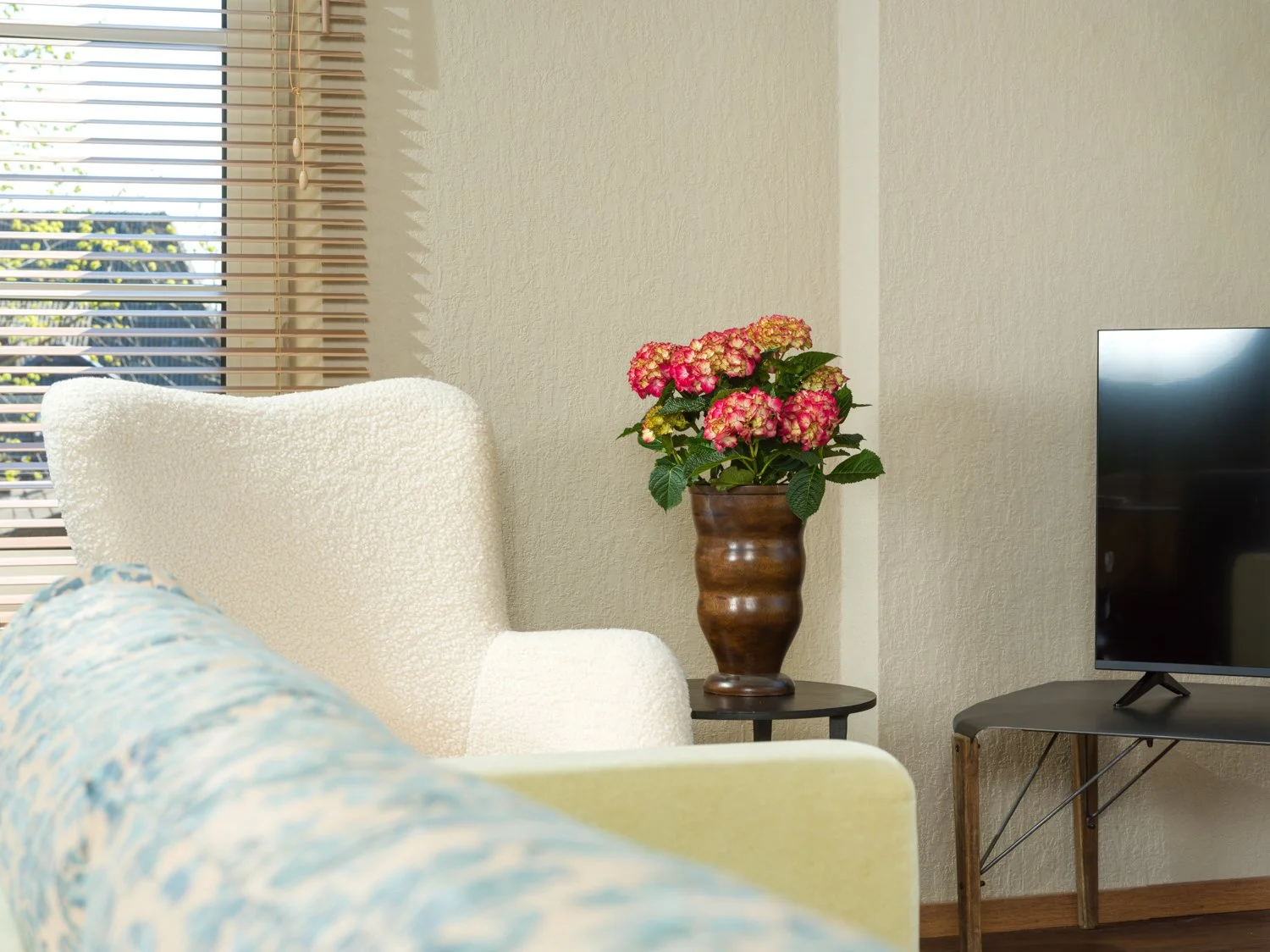 Living room with window blinds, a white armchair, a side table with a pink hydrangea plant in a brown vase, and a flat-screen TV on a stand.