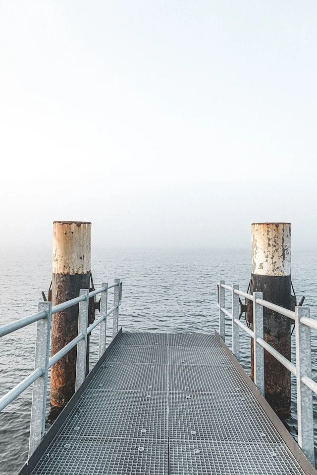 A metal dock extending over a body of water, flanked by two weathered wooden pilings, with a foggy sky in the background.