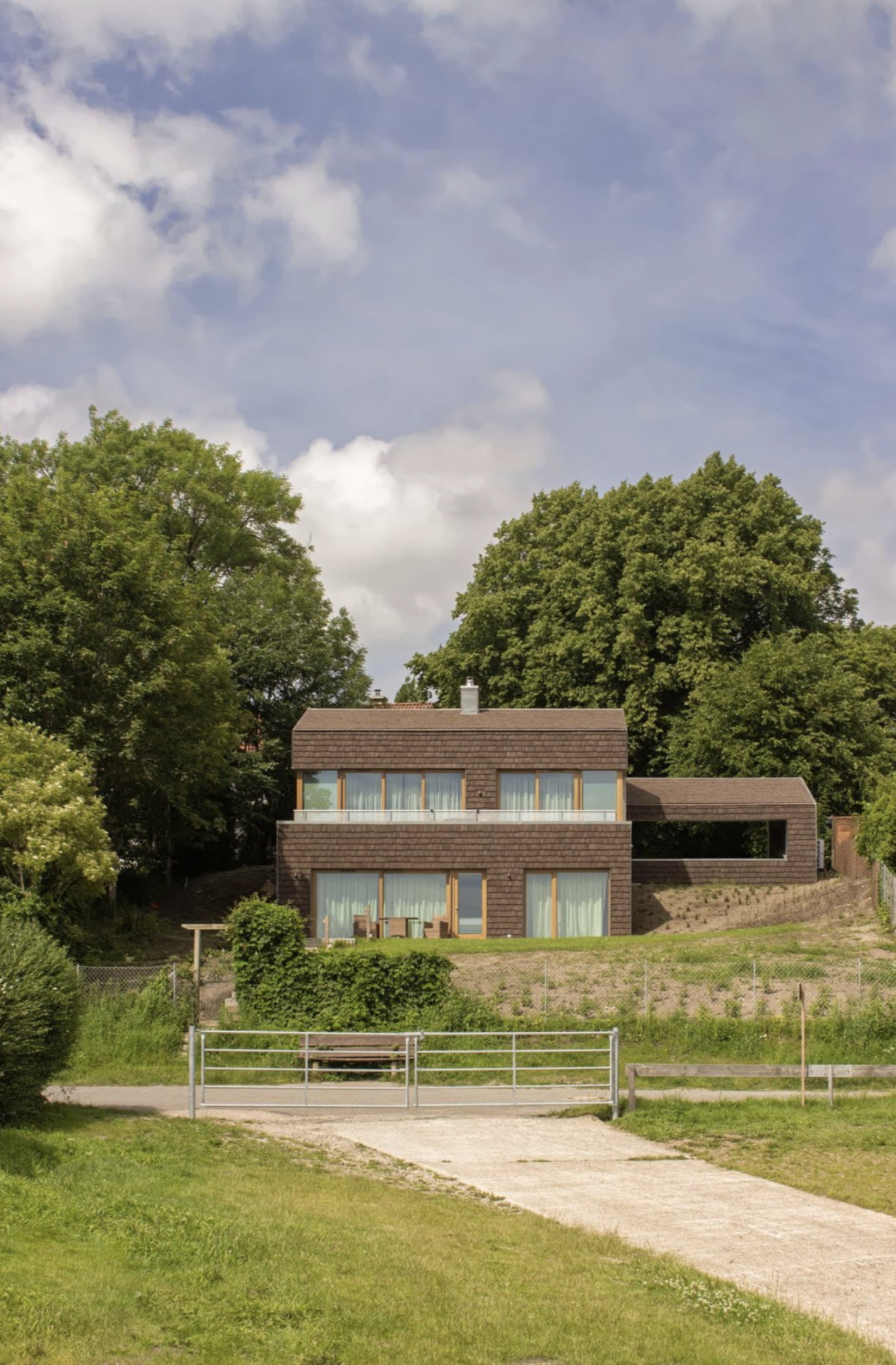A modern multi-story house with large windows, surrounded by trees and a grassy yard, under a partly cloudy sky.