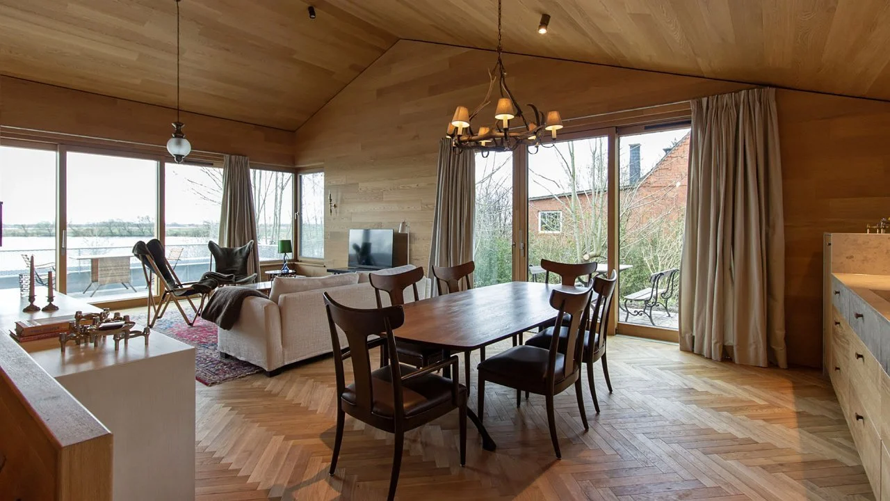 Living room with wooden walls and ceiling, featuring a dining table with six chairs, a white sofa, a TV, and large sliding glass doors leading to a balcony with outdoor seating and a view of trees and water.