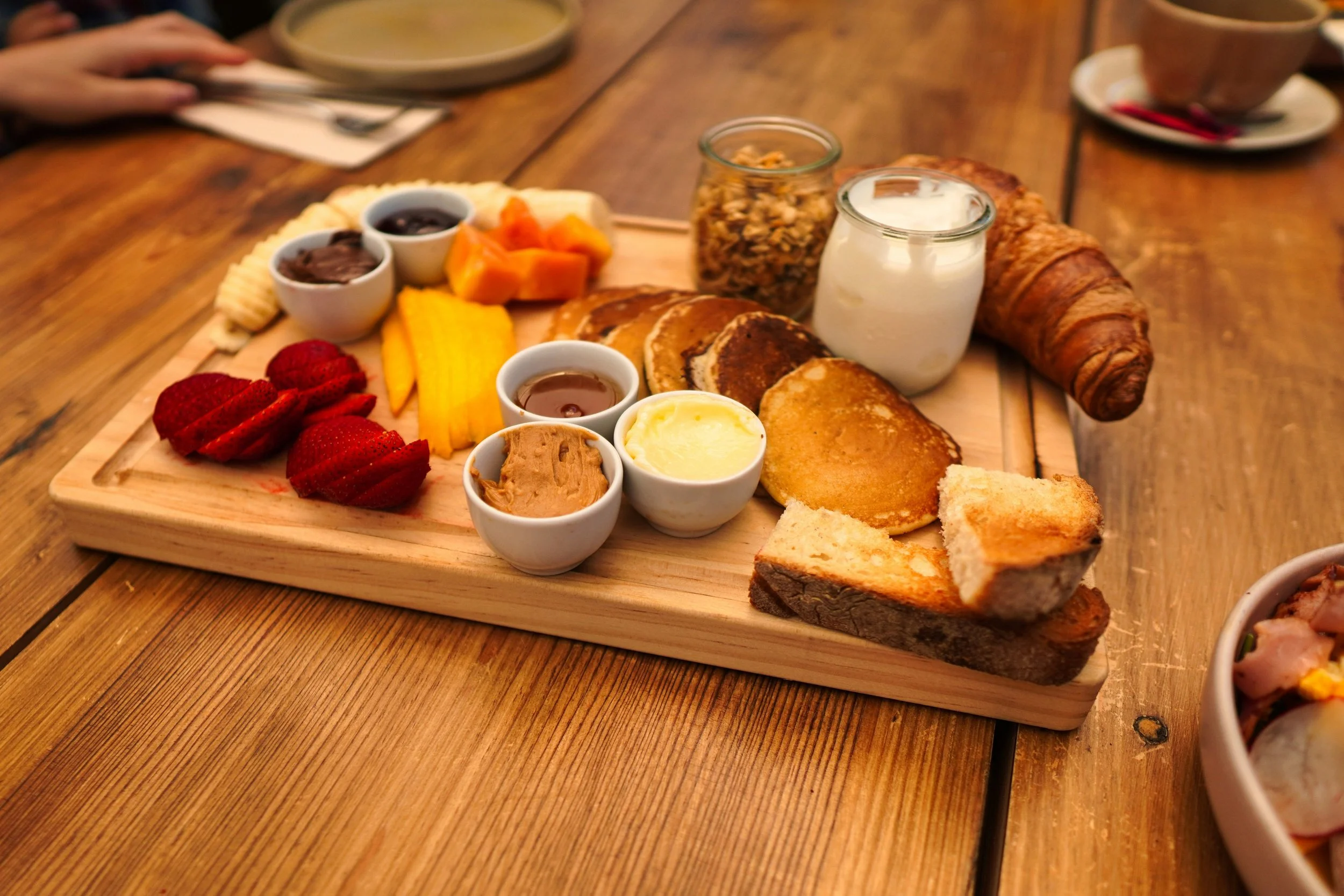 A wooden serving board with assorted breakfast items including sliced strawberries, mango slices, pancakes, croissants, bread, and small bowls of yogurt, jams, and peanut butter, on a wooden table.
