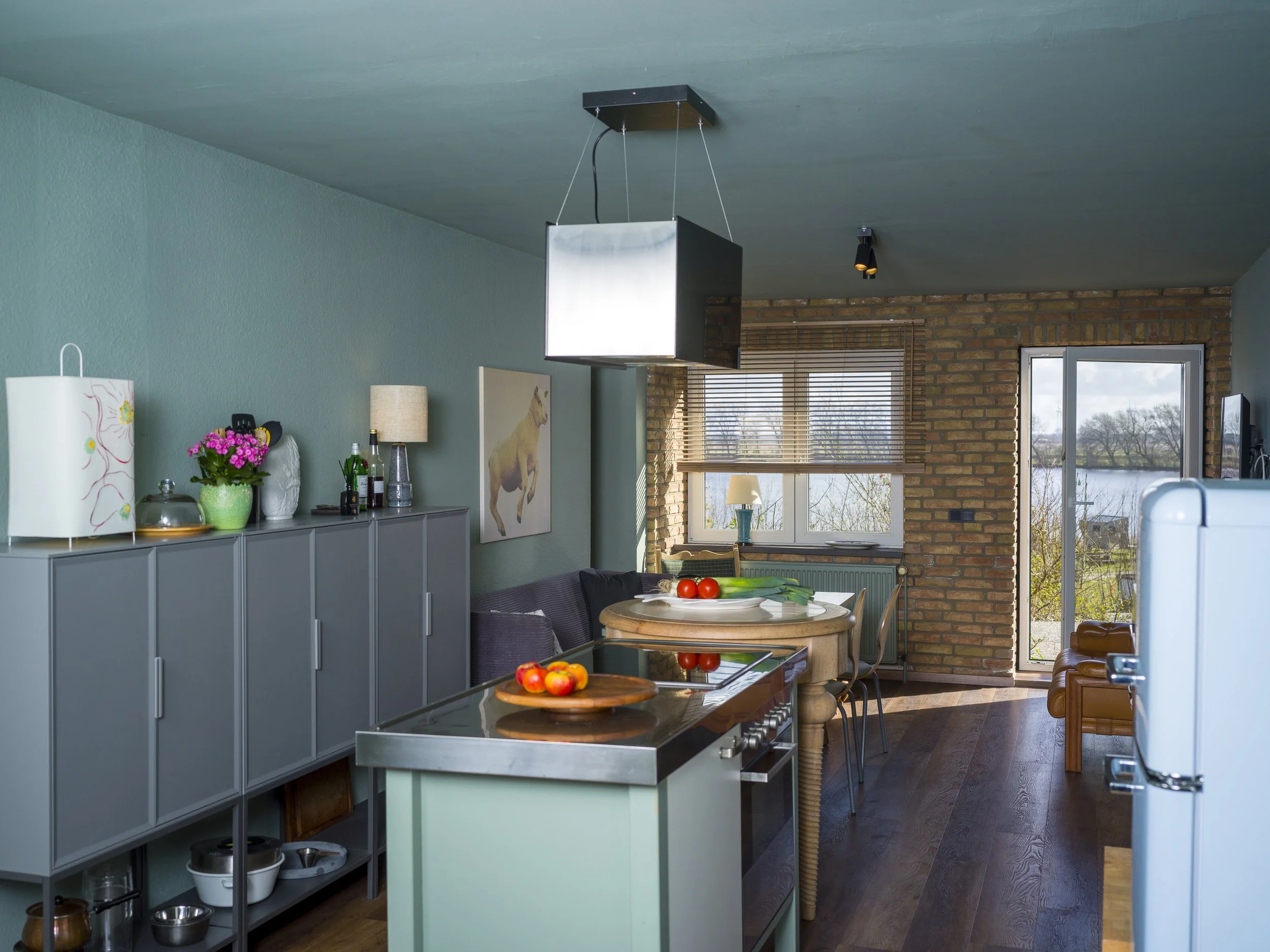 A kitchen with a blue cabinet, a wooden table with a bowl of apples, a counter with bottles and a lamp, a brick wall near a window with blinds, and ceiling lighting fixtures.