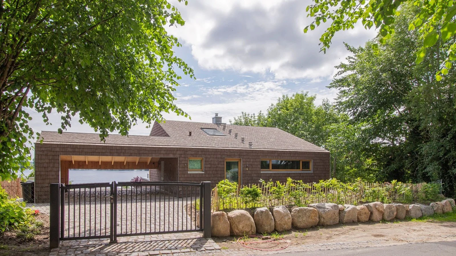 Modern house with a brown shingle exterior, situated behind a black metal gate, surrounded by large rocks and lush green trees.