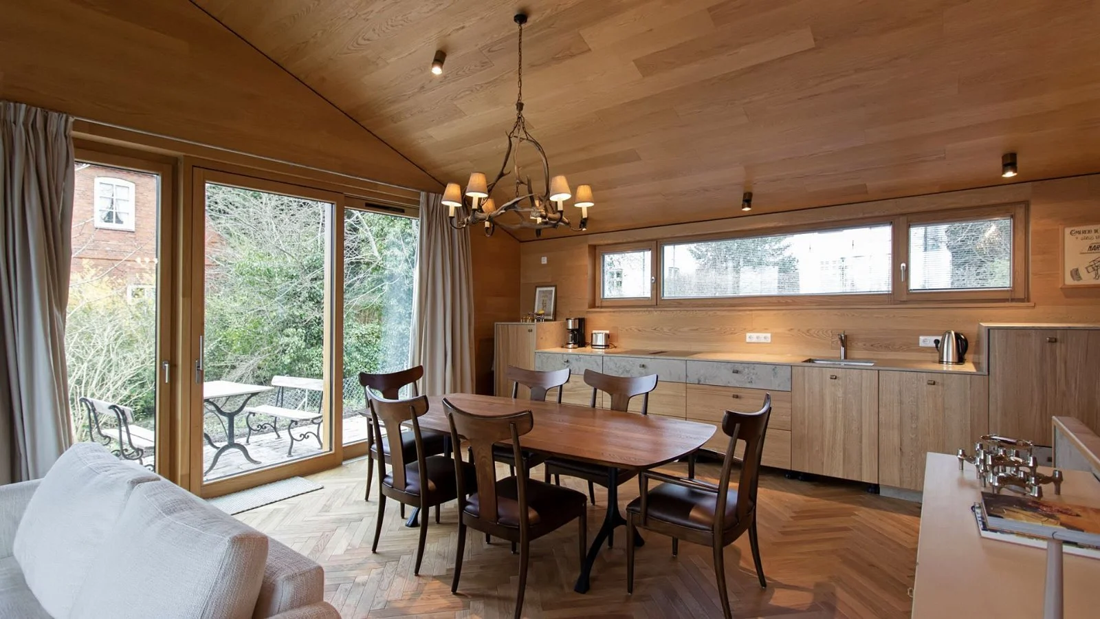 Interior of a wooden kitchen and dining area with large glass sliding door, a wooden dining table with six chairs, and a modern kitchen with countertop appliances and a long horizontal window.
