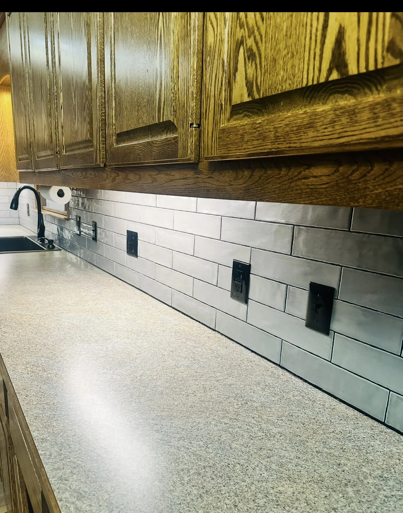 A kitchen countertop with a speckled beige surface, a black sink, and a black faucet, with wooden cabinets above and a gray tile backsplash behind.