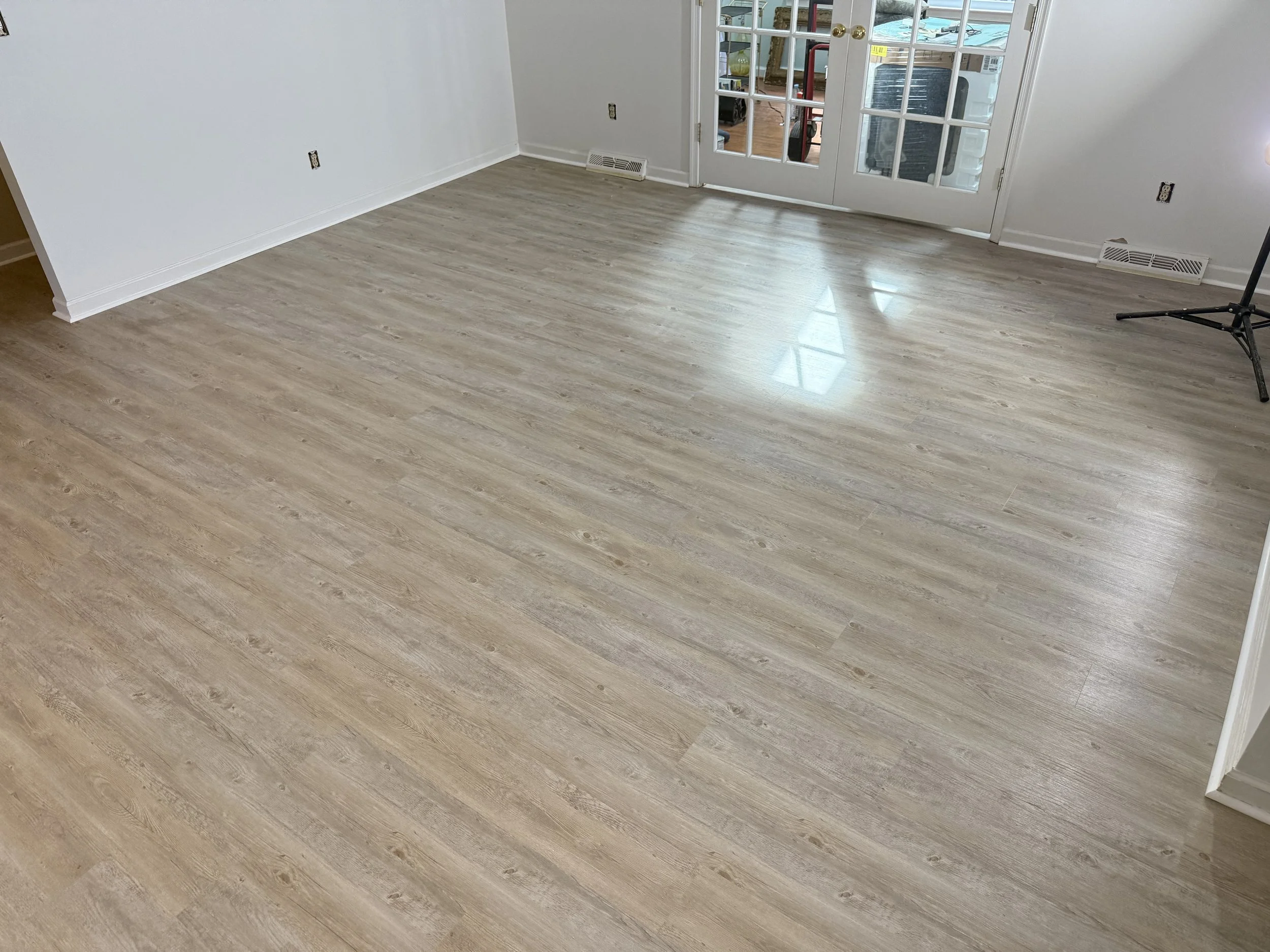 Empty room with light wood flooring, white walls, and glass double doors leading to an outdoor area. A small black tripod stand is on the right side.