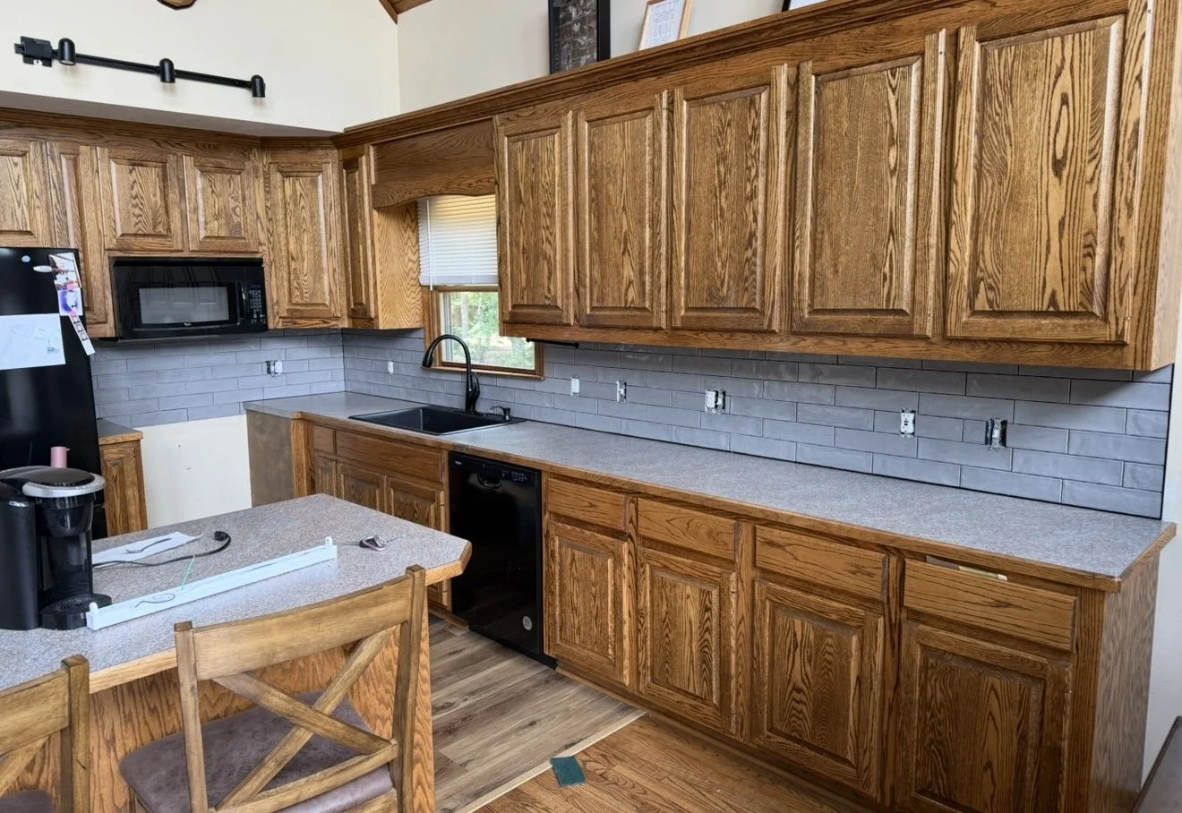 Kitchen with wooden cabinets, a black microwave, a window above the sink, gray tile backsplash, and a black dishwasher. There is a small countertop area with items including a coffee maker, a strip, and a pair of keys. The floor is wooden, and there 