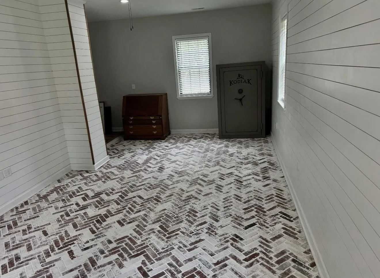 Empty room with white shiplap walls, a patterned brick floor, two windows with blinds, an old wooden writing desk, and a gray safe labeled 'KODAK'.