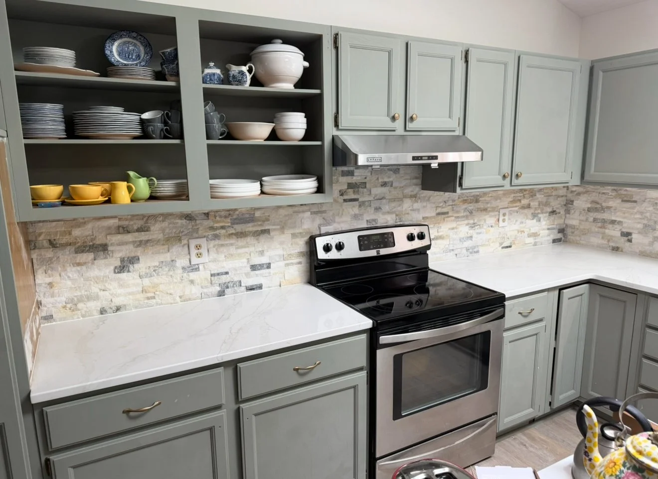 Kitchen with light gray cabinets, open shelves with dishes and cups, stainless steel stove, white countertops, stone backsplash, and some electrical outlets.
