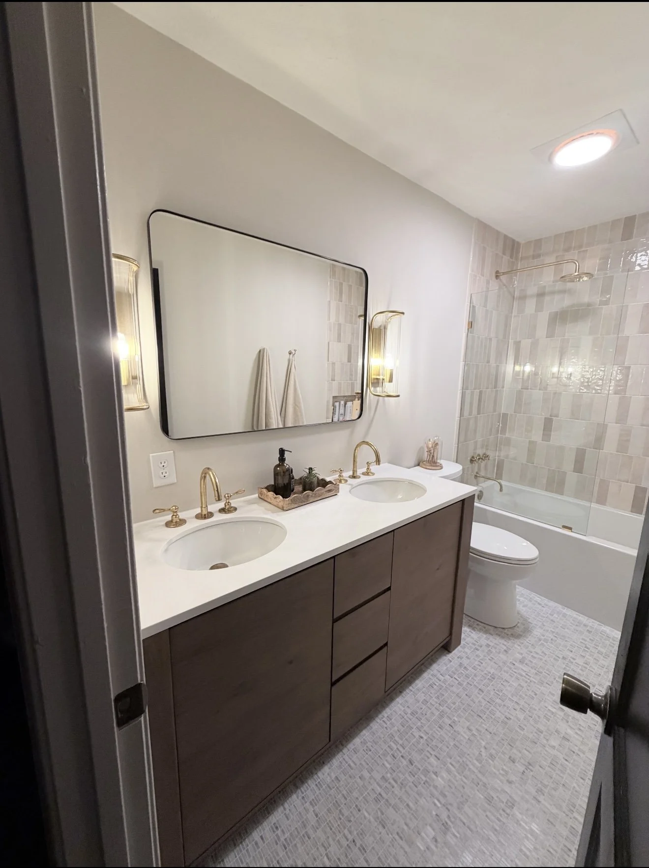 A modern bathroom with a double vanity featuring a white countertop, two oval sinks, brass faucets, and a wooden cabinet below. Above the vanity is a large rectangular mirror with black framing and wall-mounted brass light fixtures on each side. To t