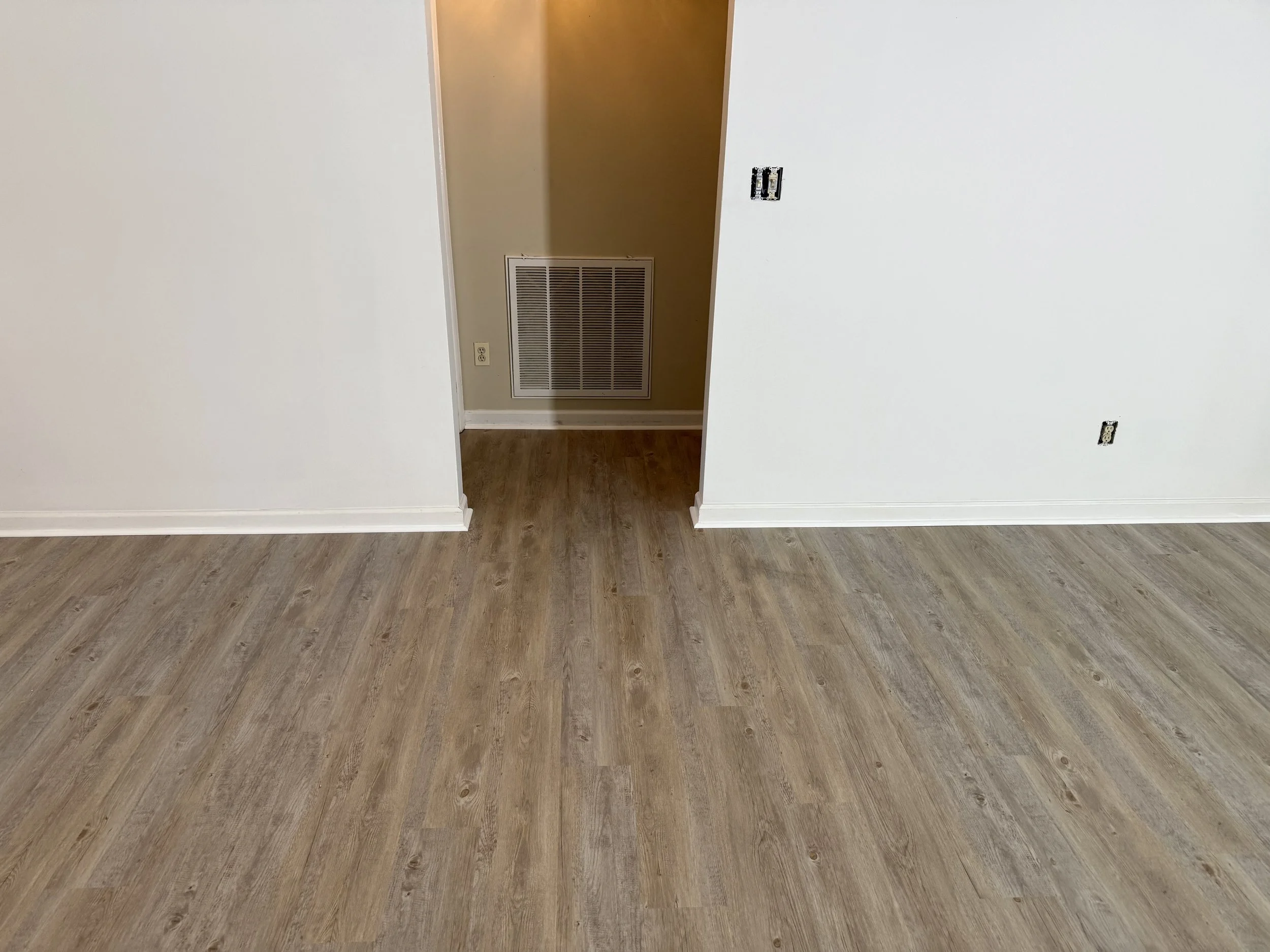 Empty room with walls painted white, unfinished electrical outlets, a vent in the back wall, and hardwood flooring.