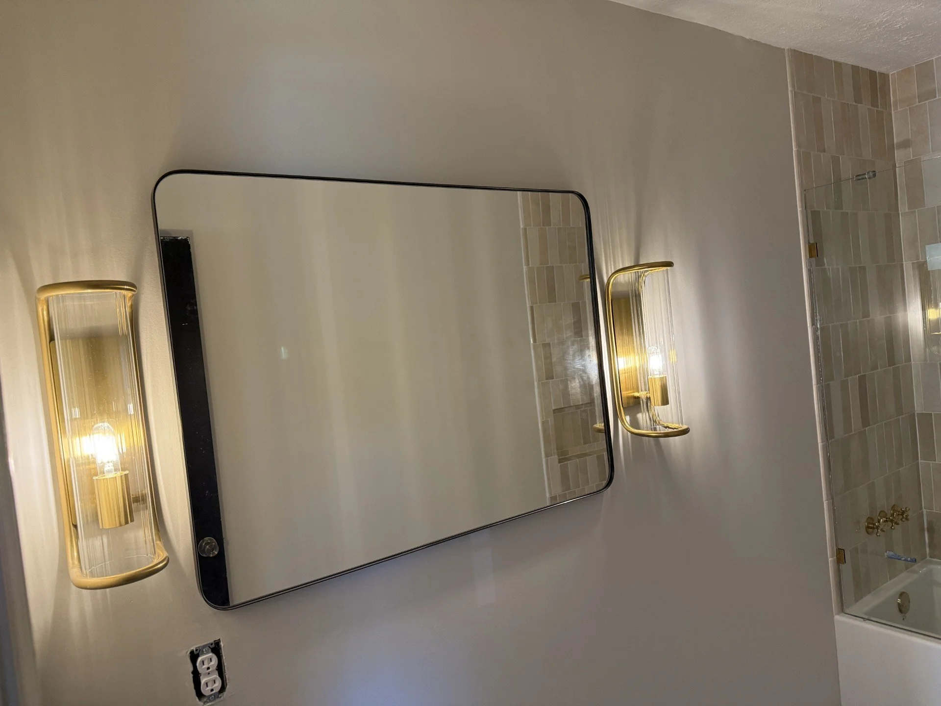A bathroom vanity mirror with two gold-colored light fixtures on either side, a beige tiled wall, and an electrical outlet below the mirror.
