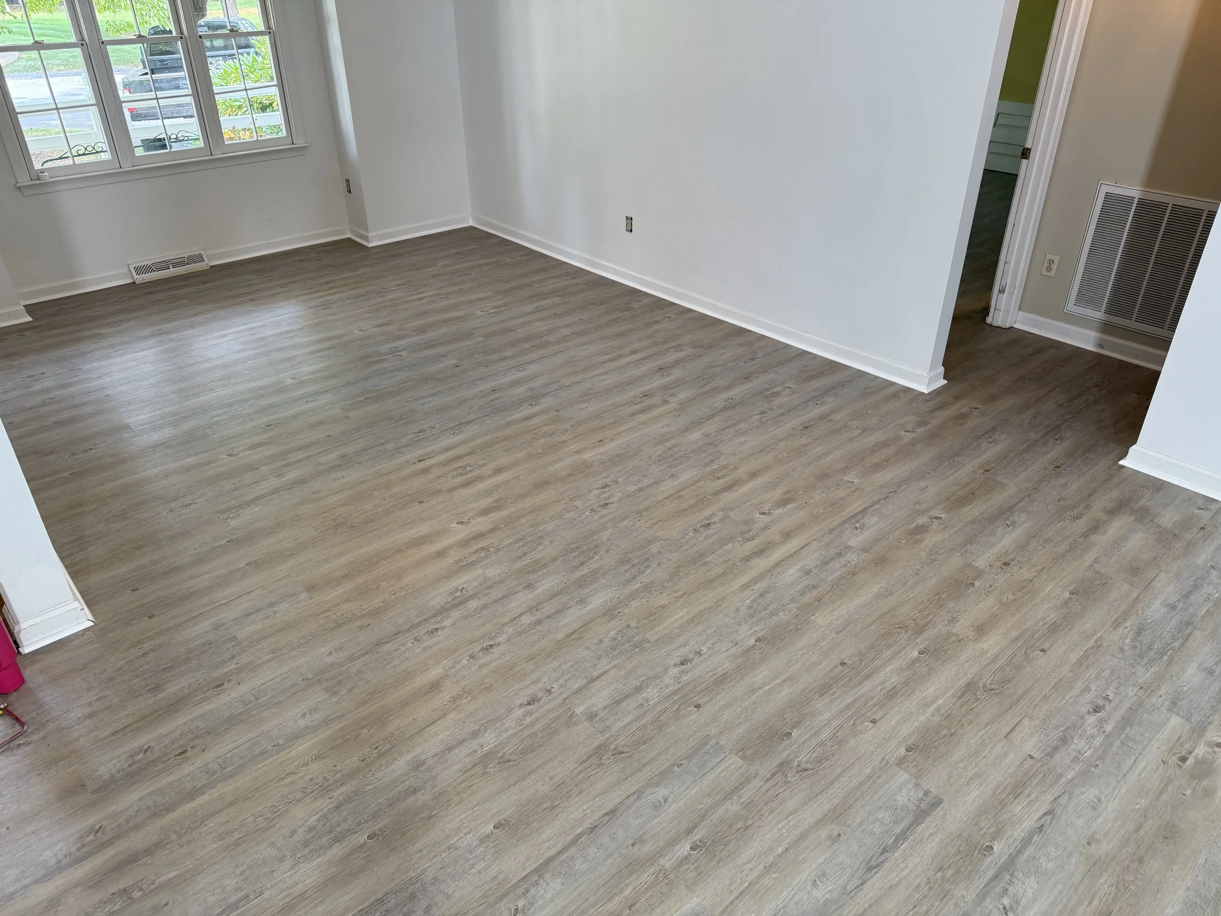 Empty room with beige walls, wooden flooring, a window with multiple panes, a vent on the floor near the window, and a door leading to another room.