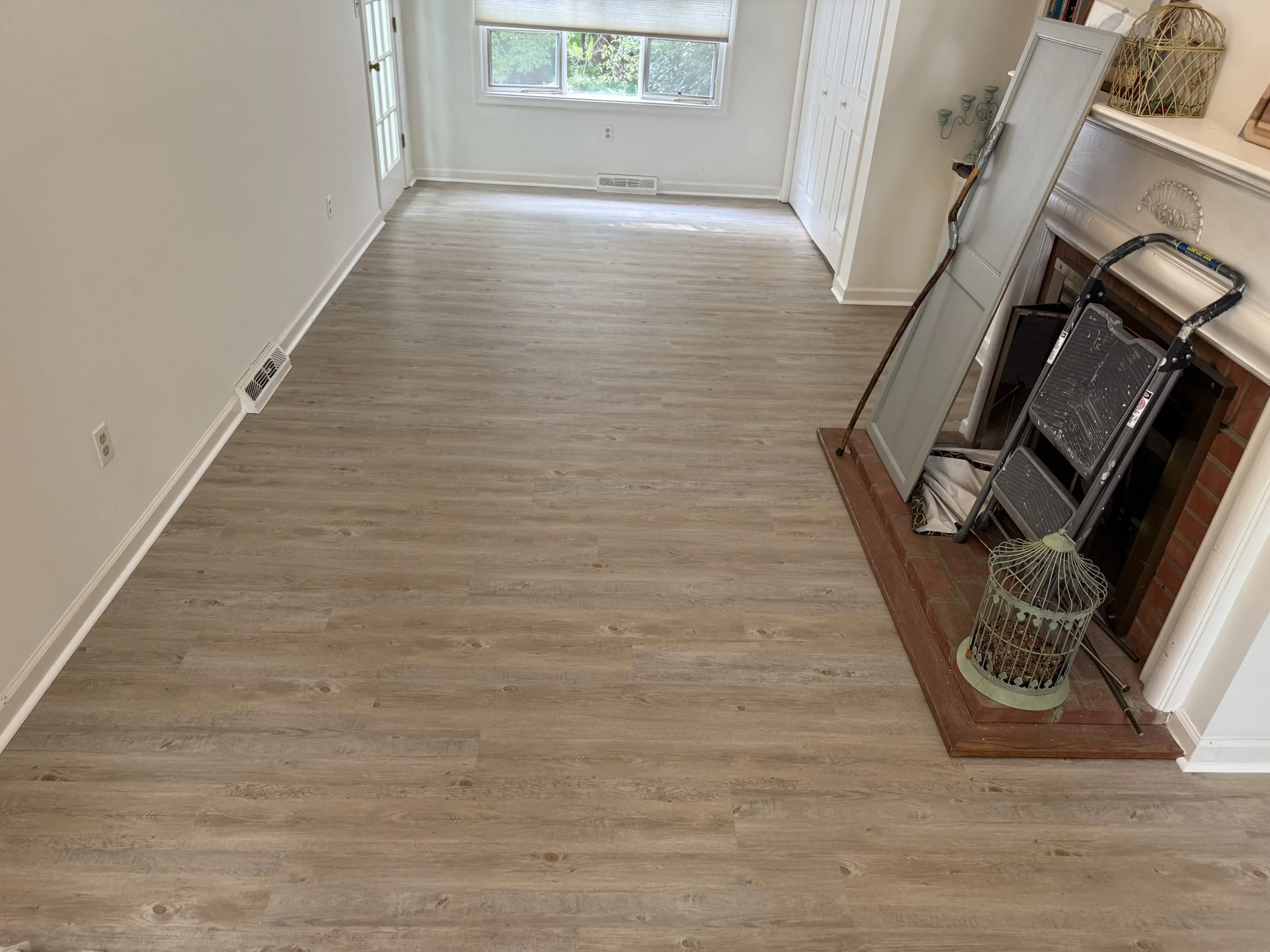 Empty living room with light-colored wood floors, white walls, large window, and closed white door. A fireplace is on the right, with a foldable ladder and various decorative items on the mantel.