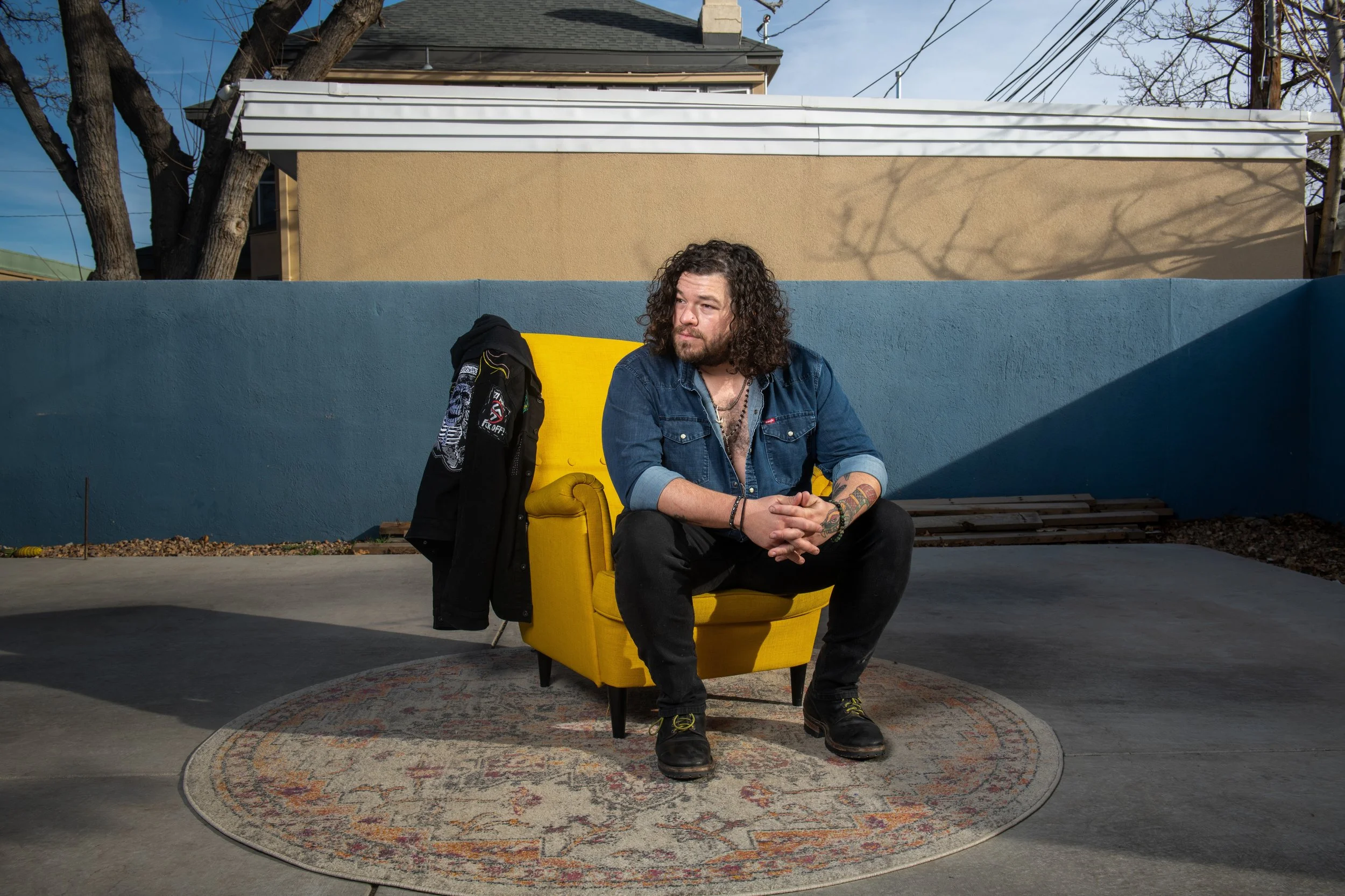 A man with curly hair and tattoos sitting on a yellow chair outdoors on a round patterned rug, with a blue wall, beige building, and trees in the background.
