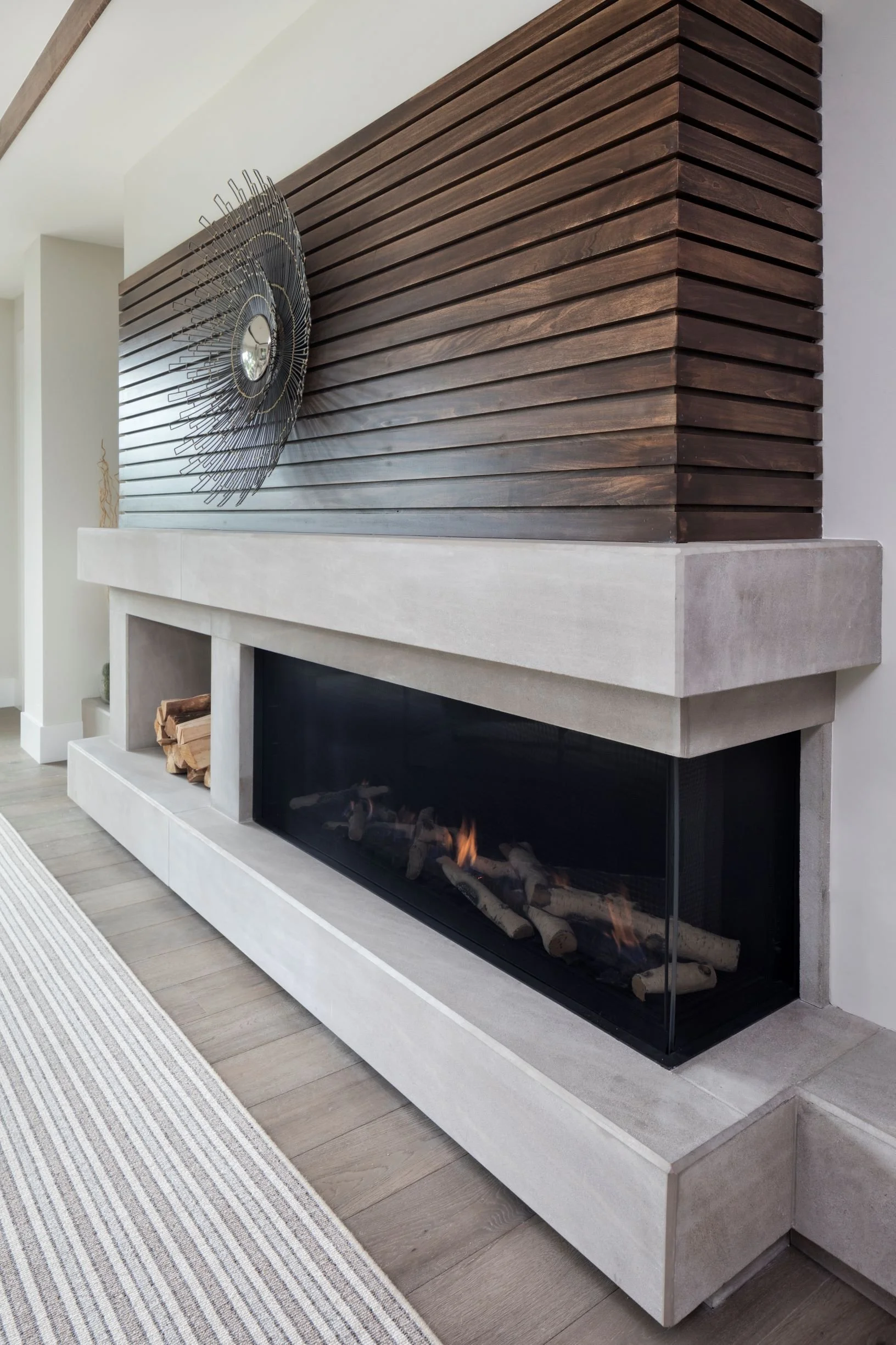 Modern fireplace with a white concrete mantel and firewood inside, above a wooden slatted wall with a decorative mirror, in a contemporary living room with a striped rug and wood flooring.