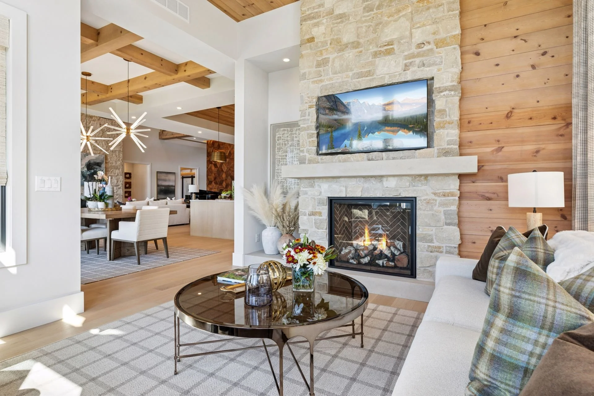 A modern living room with a stone fireplace and a mounted flat-screen TV showing a mountain lake scene. The room features a white sofa with plaid pillows, a round coffee table with decorative bottles and a flower arrangement, and a patterned rug. The walls combine white paint and wood paneling, with a ceiling of exposed wooden beams.