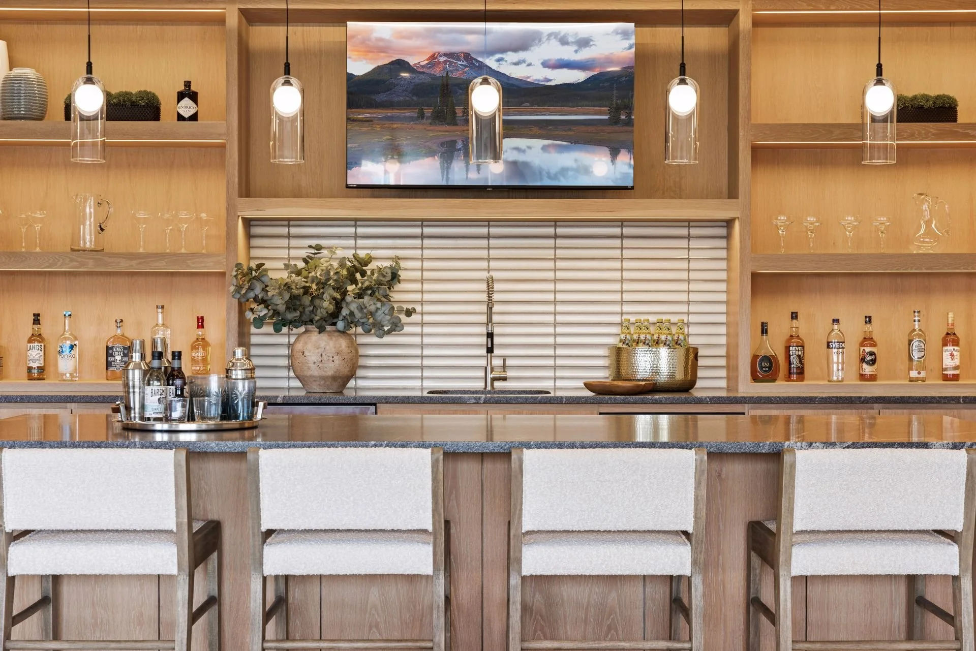 A wooden bar countertop with white chairs, bottles of alcohol, glasses, a tray, a vase with greenery, a gold ice bucket with bottles, and a mounted landscape photograph of a mountain and lake above the bar with hanging lights.