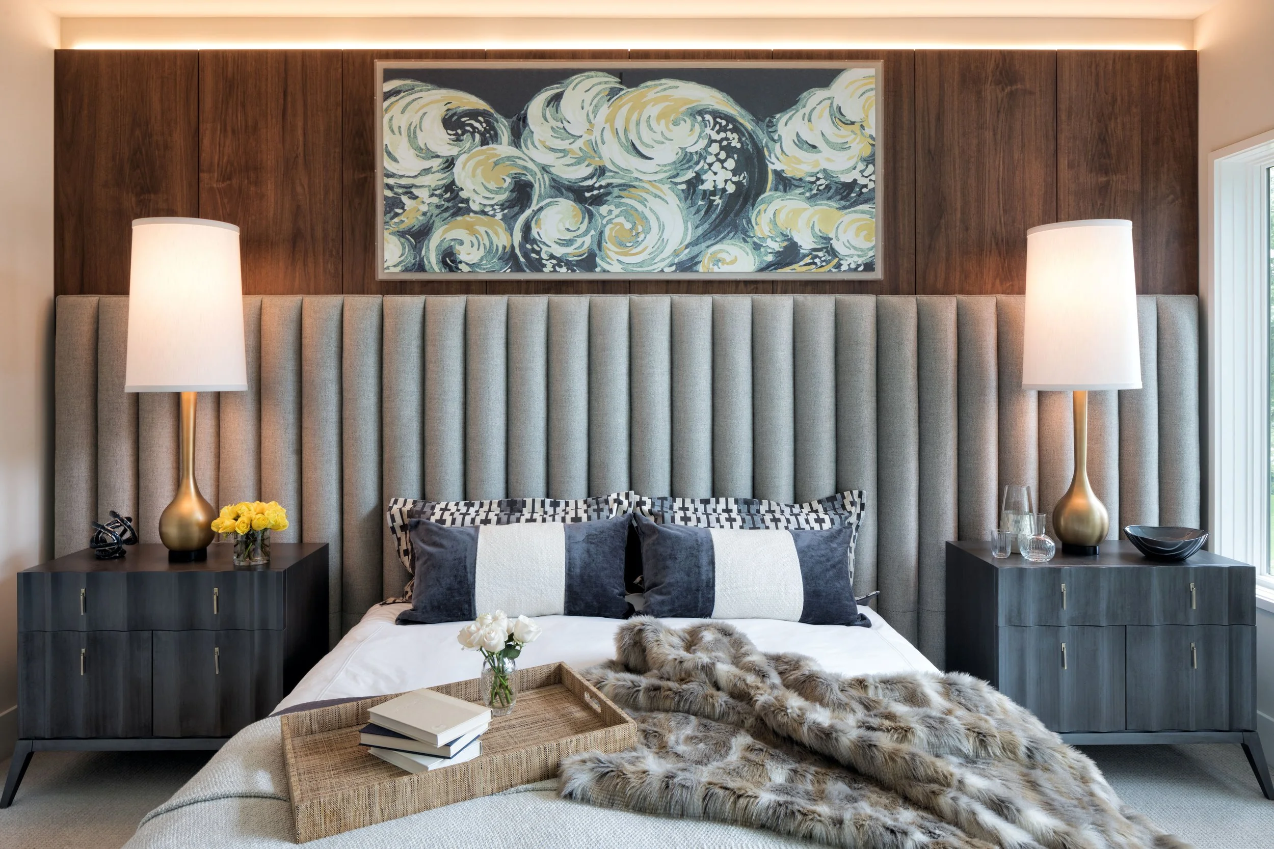 A modern bedroom with a large channel tufted headboard with custom walnut panels behind, two nightstands with lamps, a painting of ocean waves above the headboard, and a bed with black and white pillows, a tray of books, and a fur blanket.