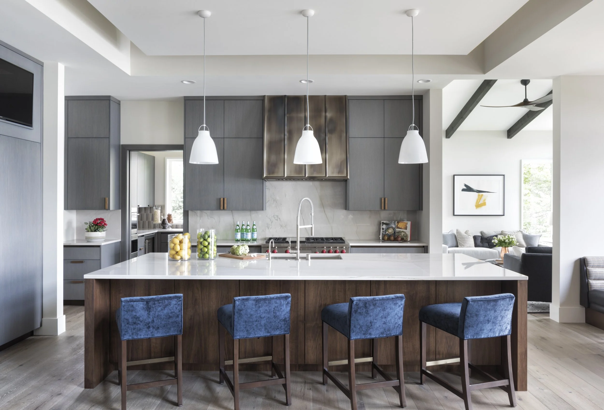 Modern kitchen with gray cabinets, white island with wood base, three white pendant lights, and blue upholstered chairs. Decor includes framed art, potted plant, and bowls of fruit.