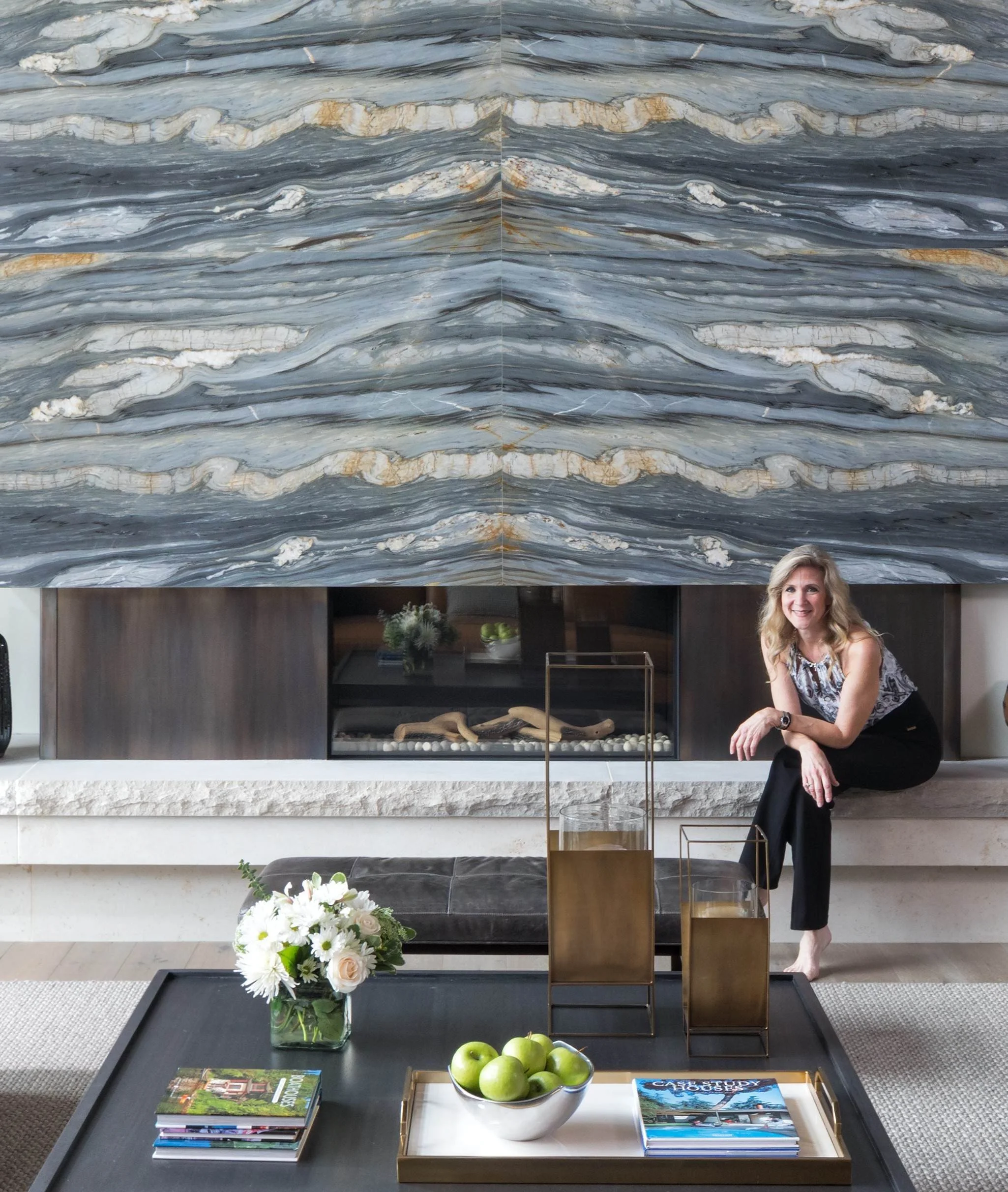 Living room with large marble fireplace, woman sitting on the carpeted floor, floral arrangement and green apples on coffee table, books, and greenery in the background.