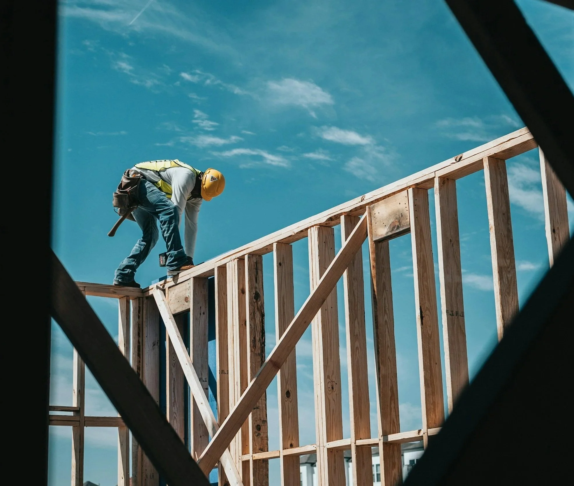 A construction worker wearing a yellow safety helmet and a reflective vest working on a wooden framework at a building site under a partly cloudy sky.