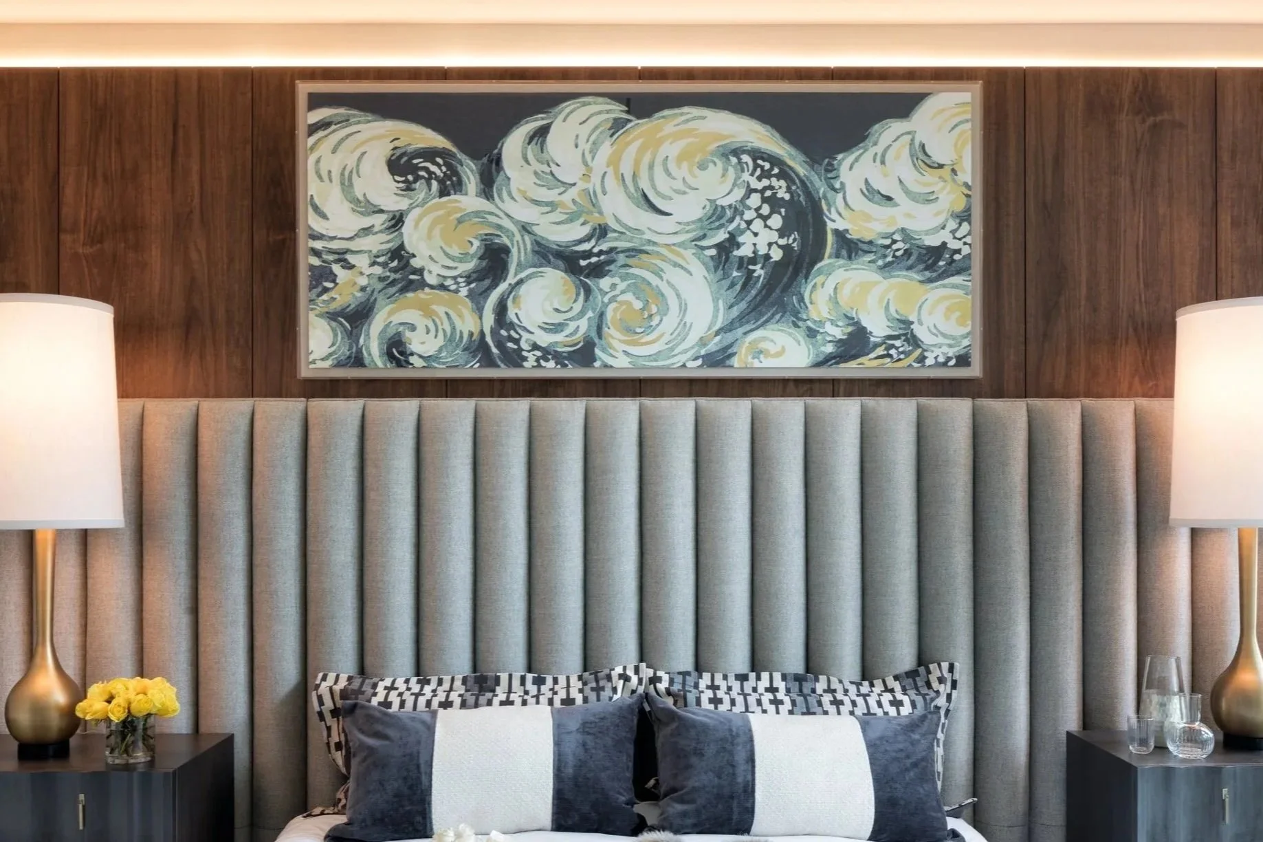 A modern bedroom with a light beige upholstered bed, dark throw pillows, and a patterned accent pillow, flanked by side tables with lamps and decorative items, and a large framed print of a swirling wave painting on a wooden-paneled wall.
