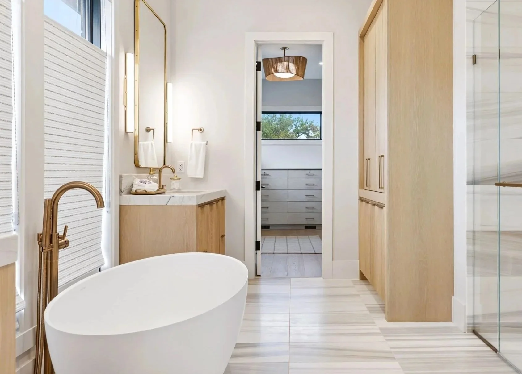 Modern bathroom with white freestanding bathtub, wooden vanity with marble countertop, gold fixtures, large mirror, light-colored wood cabinetry, glass shower enclosure, and a view into a walk-in closet with drawers and greenery visible through the window.