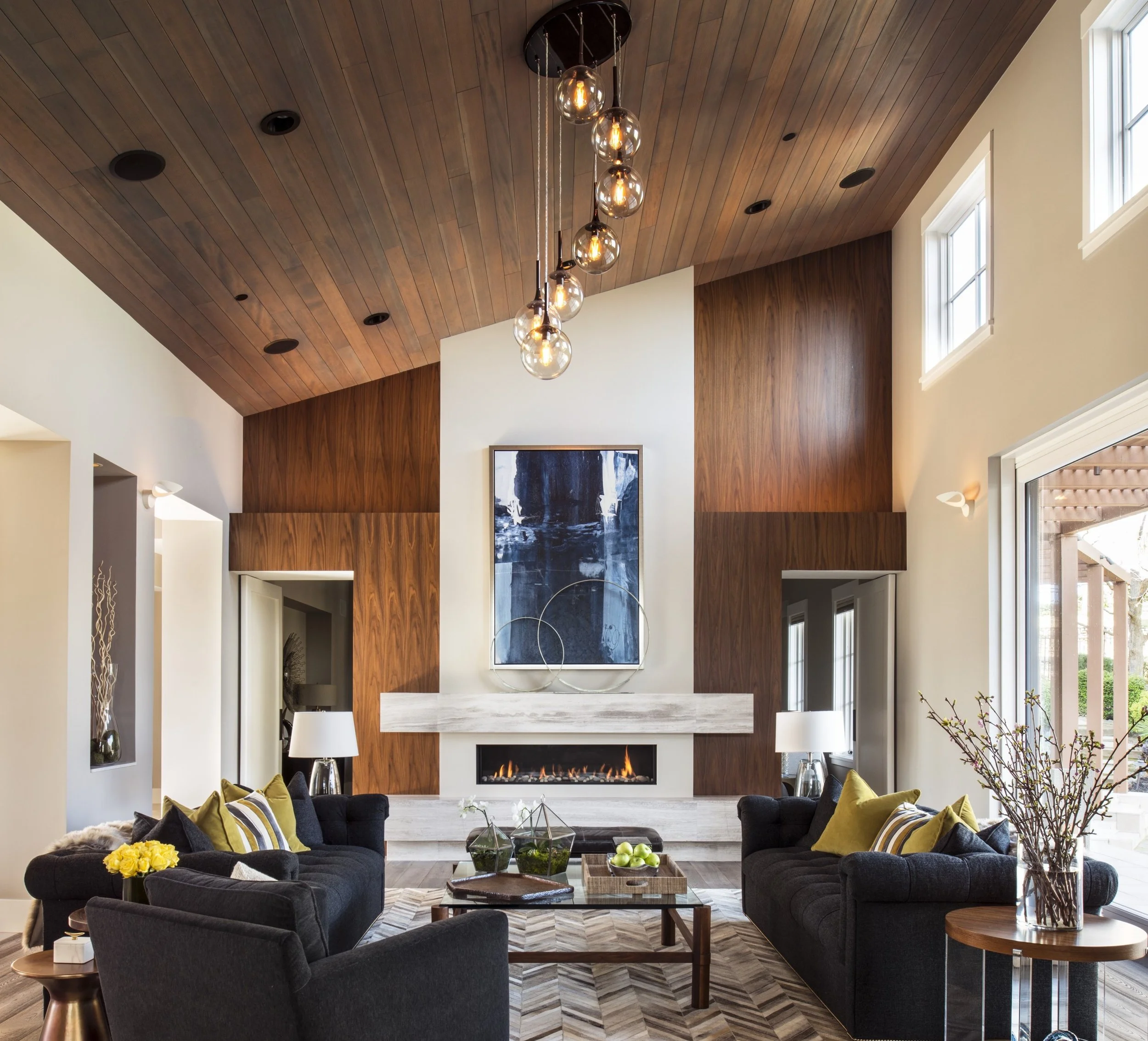 Living room with dark sofas around a wooden coffee table, a stone fireplace, a large abstract artwork on the wall, and large windows with sunlight, ceiling with wood panels, modern pendant chandelier, and decorative plants.