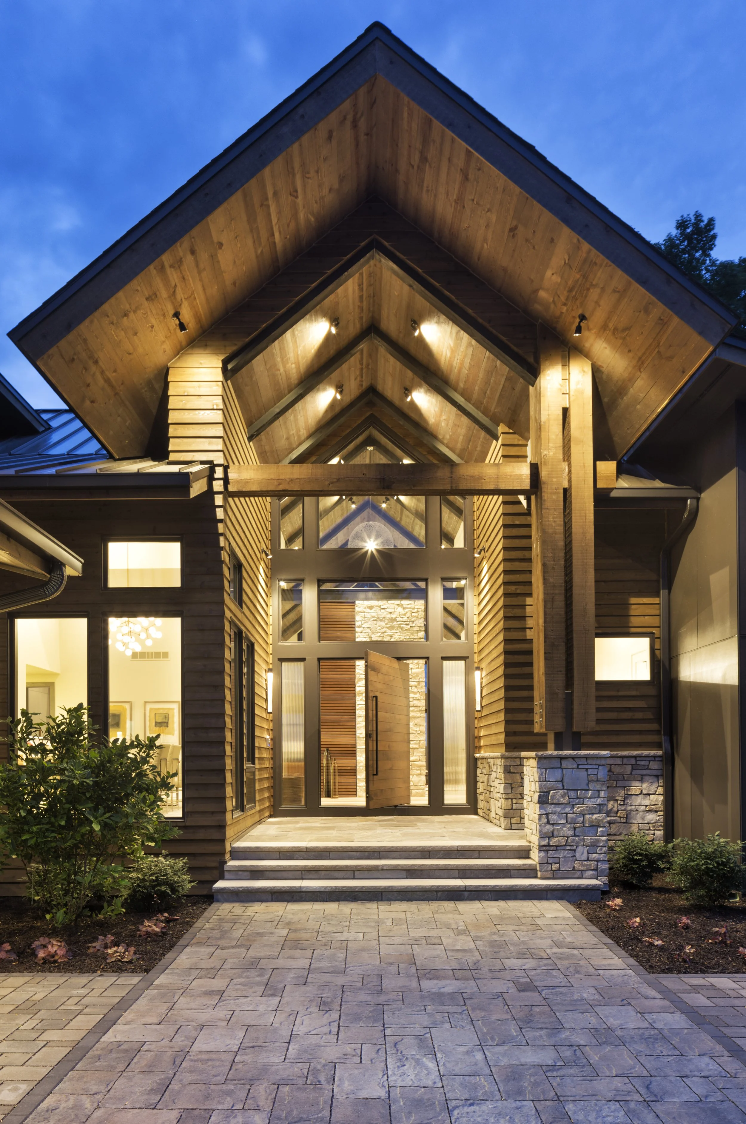 Front entrance of a modern house with a pivot walnut  door, gabled roof, stone accents, and outdoor lighting, with a paving stone walkway and landscaped garden.
