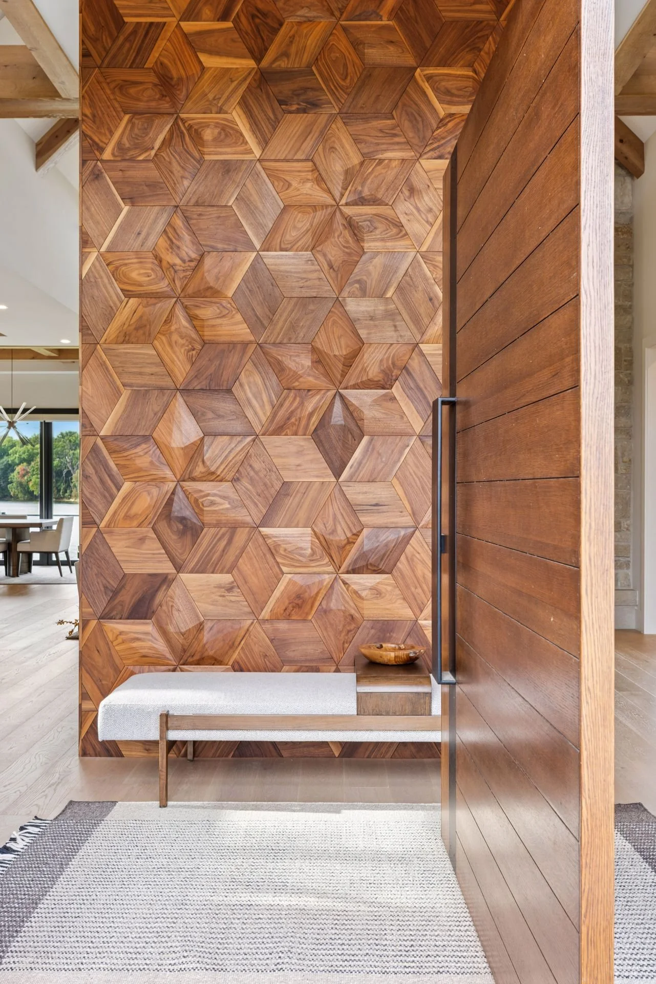 Interior view of a modern room with a custom dimensional walnut wall, a light-colored cushioned bench with wooden frame, a small side table with a wooden bowl, and a light-colored rug on the floor.
