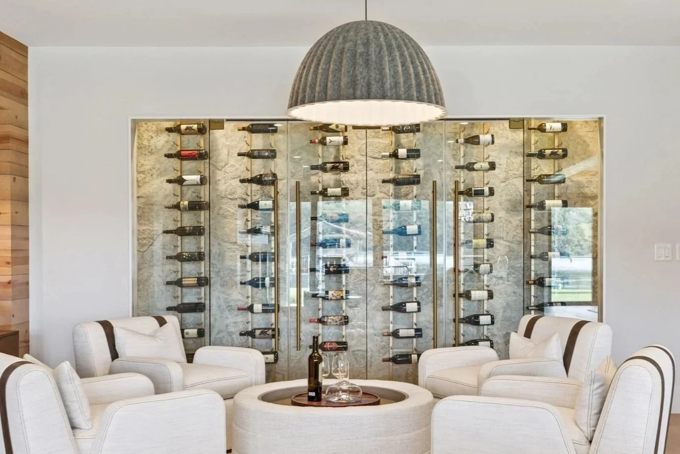 Interior of a modern wine tasting room with a circular seating area, white armchairs, and a large wall-mounted wine rack behind glass panels displaying wine bottles.
