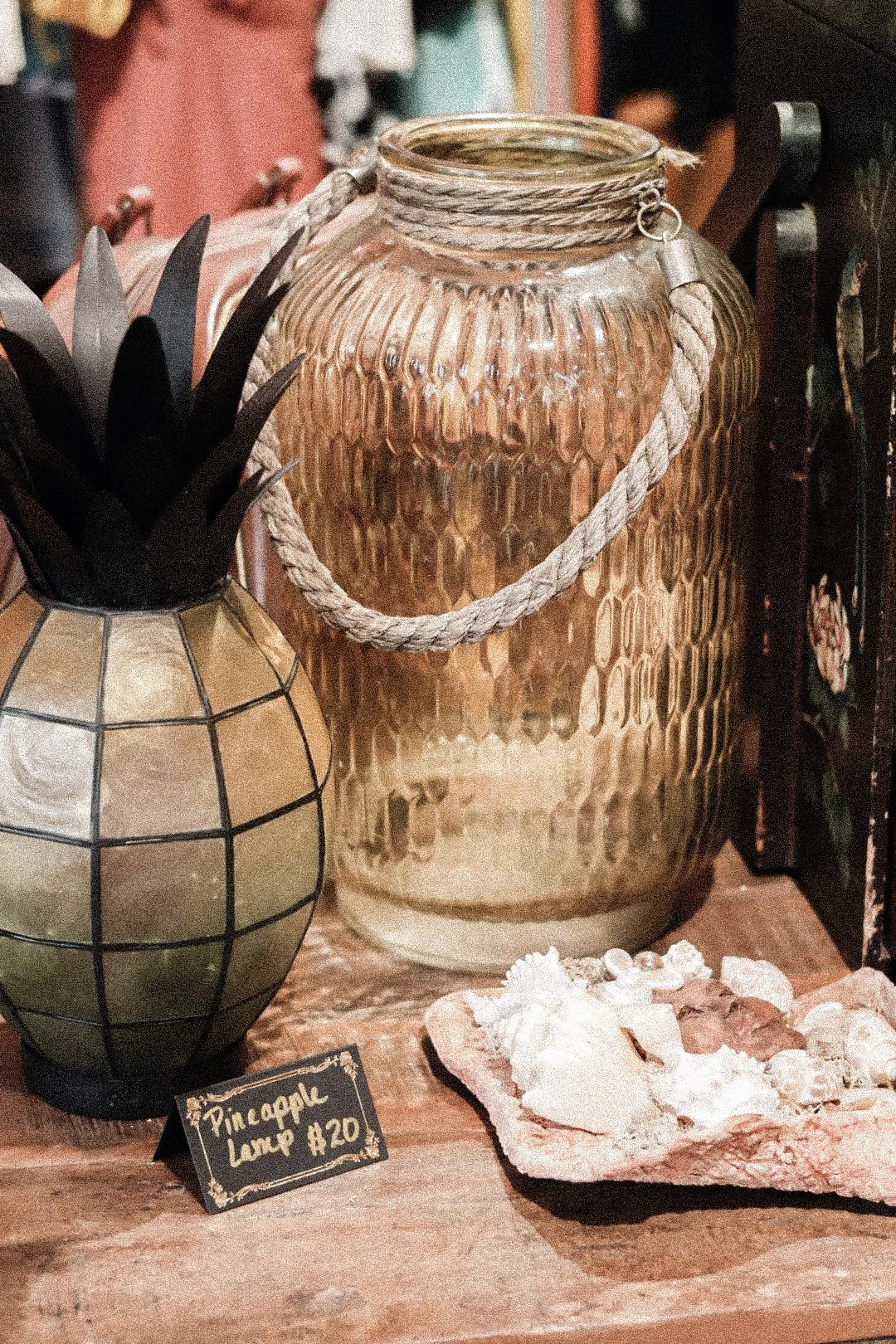 Decorative display with a large textured glass jar with a rope handle, a smaller geometric ceramic vase with plant, seashells on a wooden surface, and a small chalkboard sign reading 'Pineapple Lamp $20'.