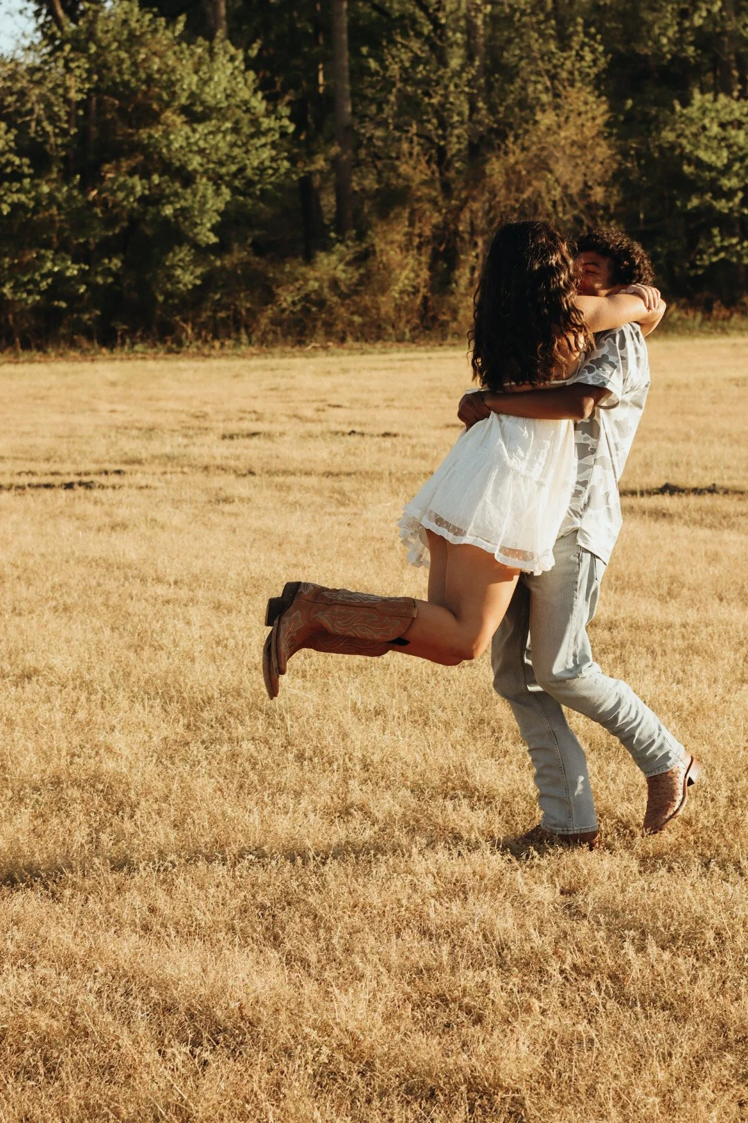 A couple is outdoors on a grassy field during sunset, with the man lifting the woman while she wraps her arms around his shoulders and they smile at each other.
