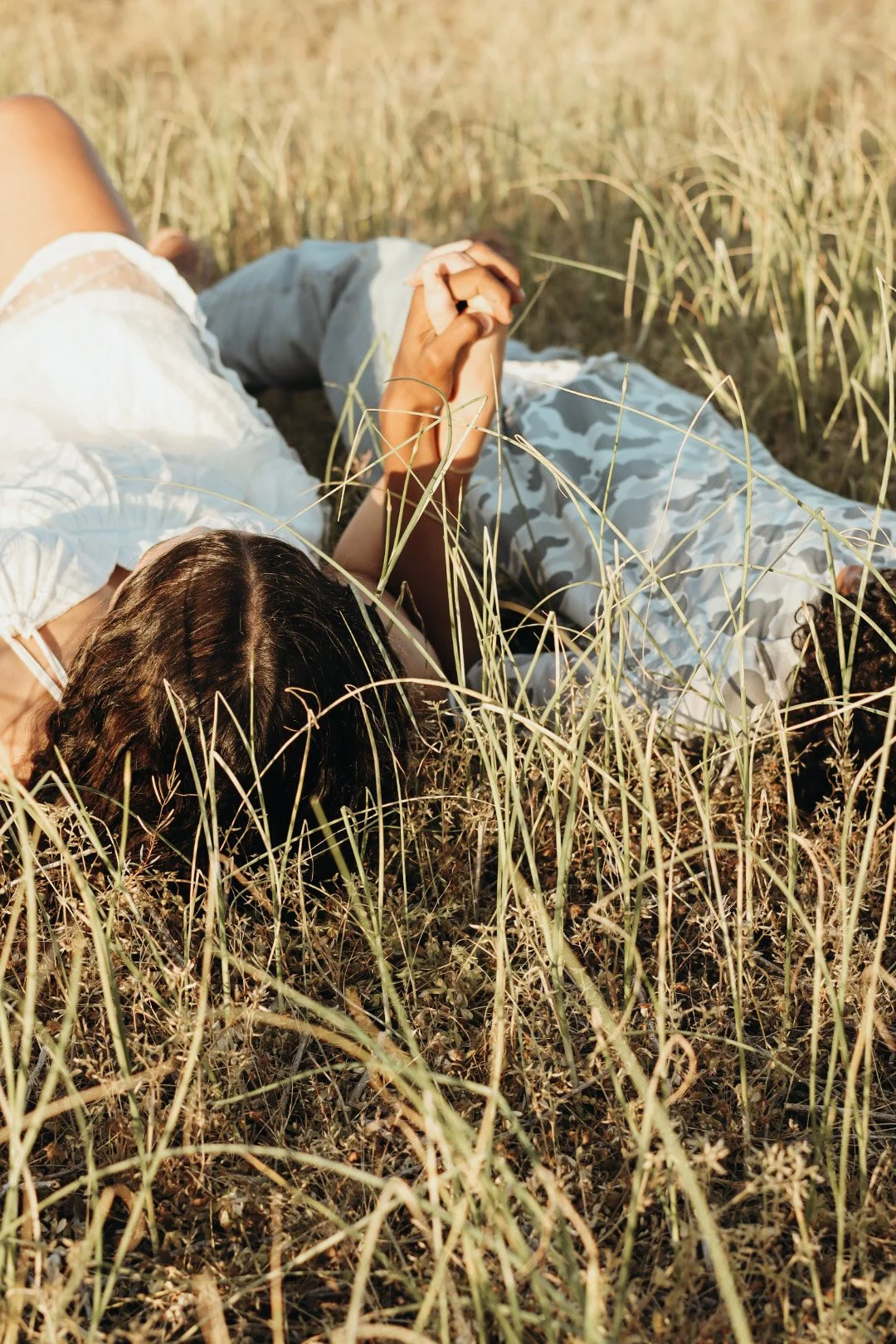 A woman lying on the grass in a field, holding hands with someone; they are surrounded by tall, dry grass and wearing casual clothing.
