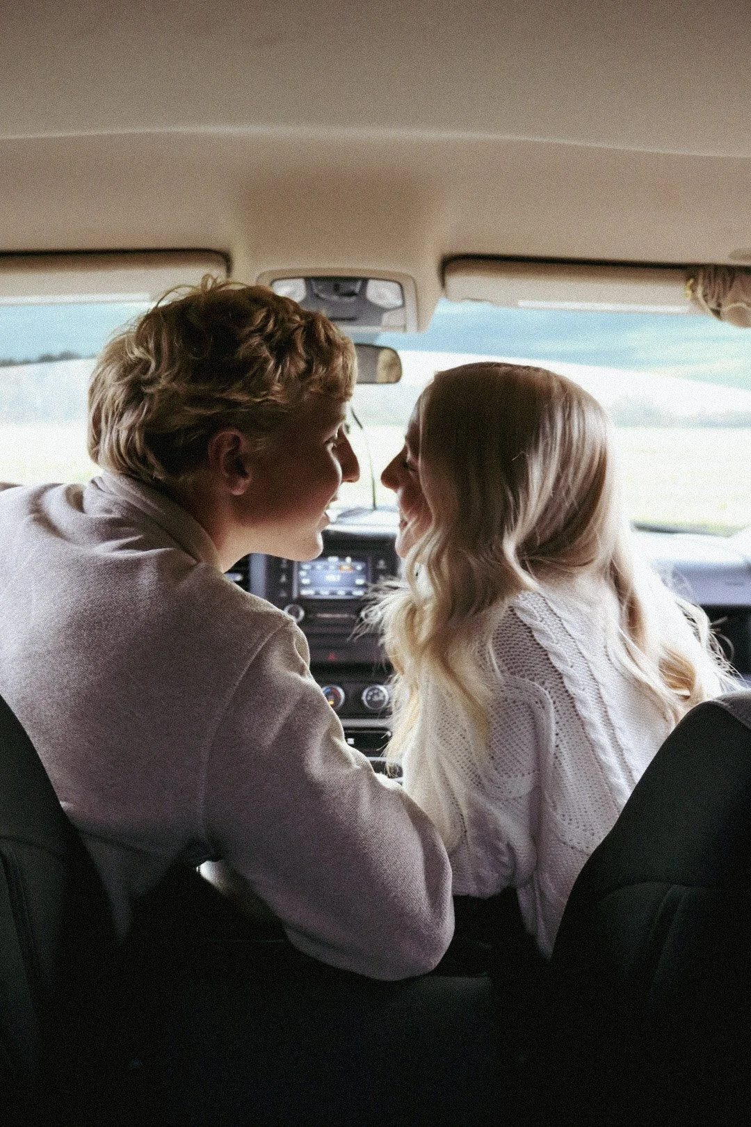 A young couple sitting in the front seat of a car, facing each other and smiling while looking into each other's eyes.