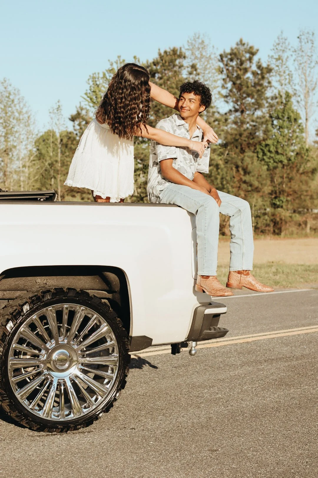 A young man and woman sitting on the edge of a white pickup truck on a rural road, with trees in the background. The woman is leaning over the man, who is smiling and looking at her, both dressed casually.