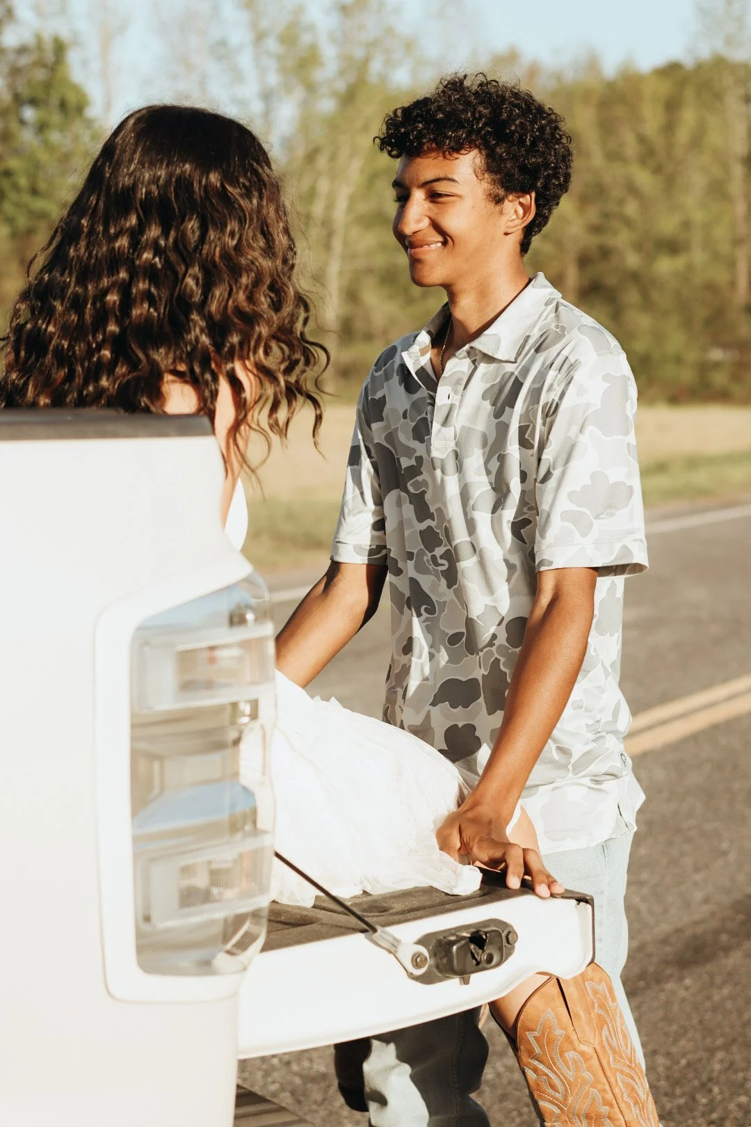 A young man and woman share a moment beside an open car trunk during daylight; the woman is in a white dress, and the man is holding her hand.
