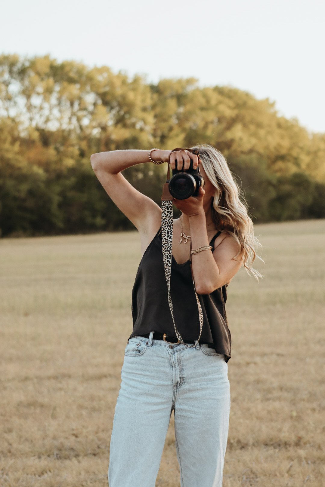 A woman with blonde wavy hair taking a photo with a camera in an open field with trees in the background during daylight.
