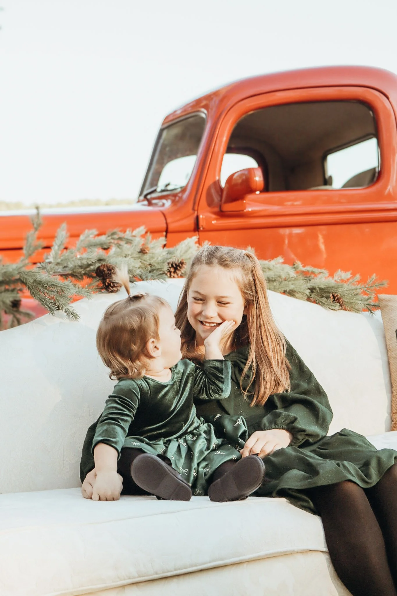 A young girl with long red hair and a smile, and a small child with blond hair sitting together on a white couch, near a decorated Christmas tree. In the background, a red vintage truck is visible, with a sprig of pine and pinecones on the vehicle.