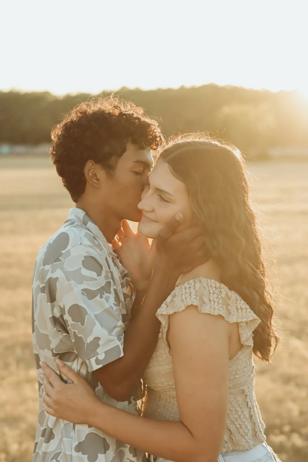 A young couple sharing an intimate moment outdoors during sunset, with the man kissing the woman's forehead and her eyes closed.