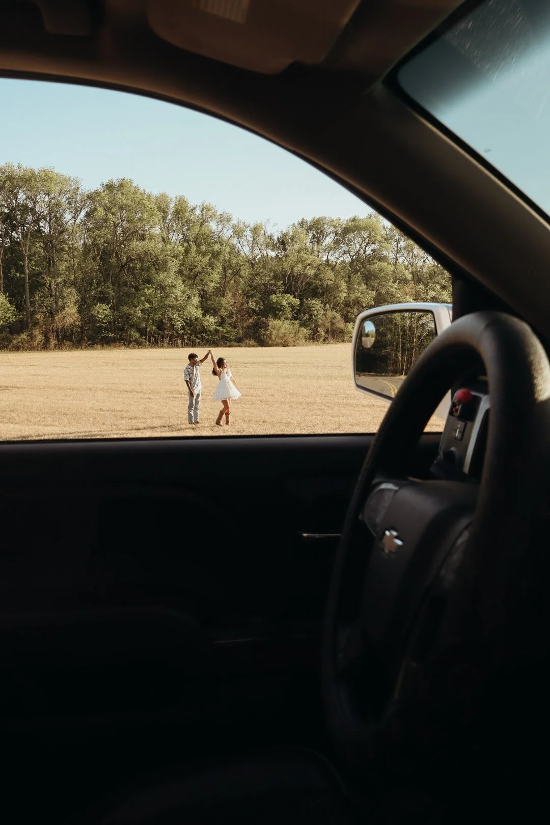 A man and woman dancing in an open field, seen from the inside of a car with the window down; the landscape includes trees in the background and a clear sky.