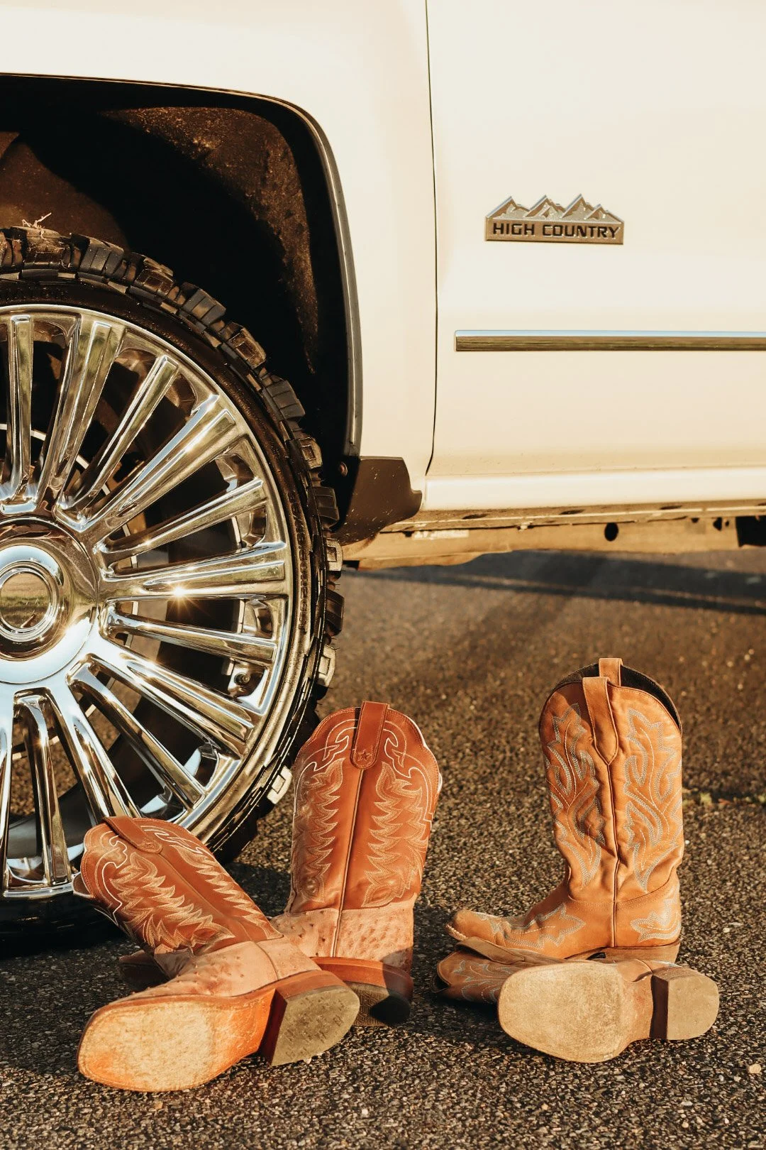 A close-up of a white vehicle, showing a chrome wheel and a "High Country" badge. Three pairs of cowboy boots, worn and scuffed, lie on the ground in front of the vehicle.