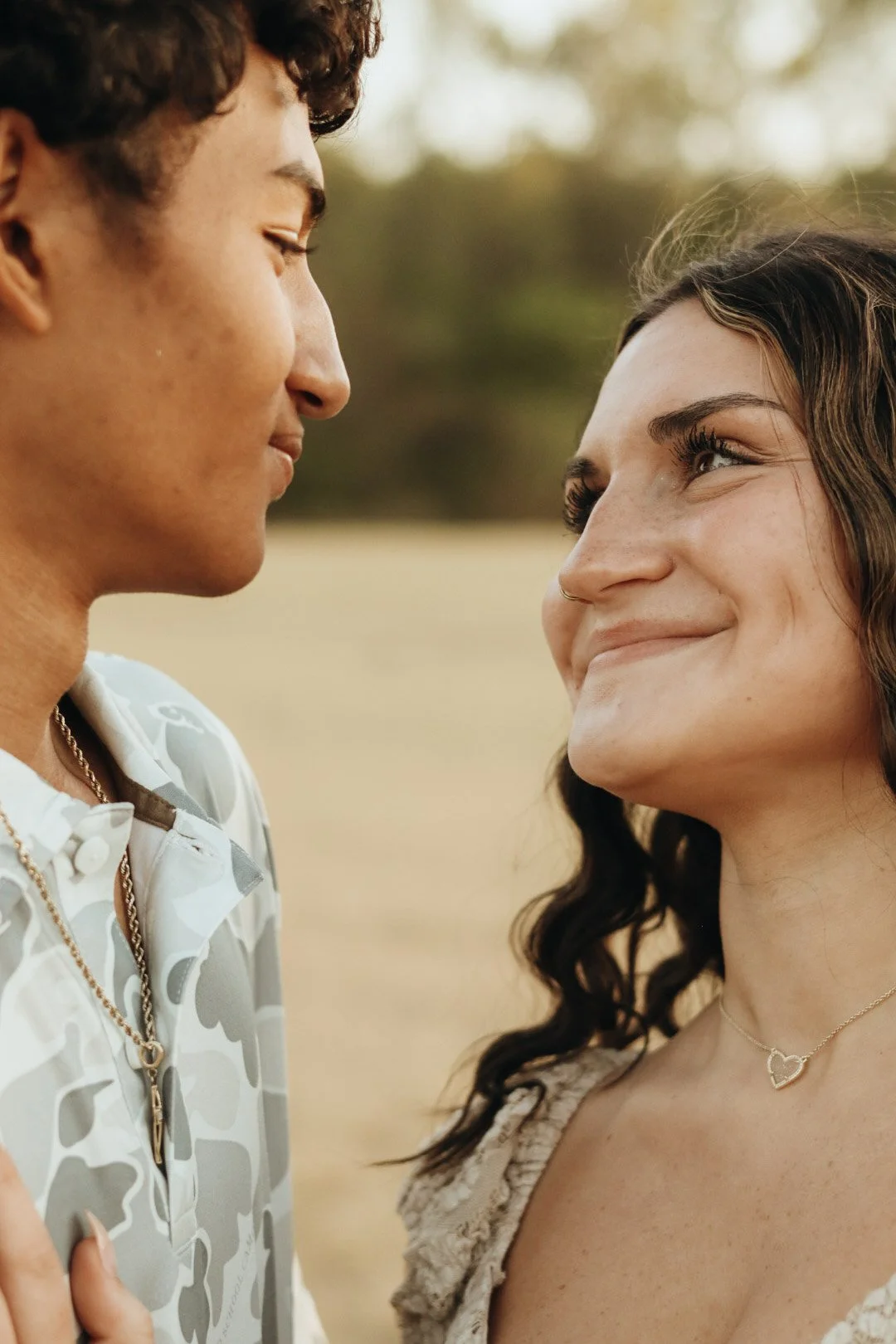 A close-up of a young couple facing each other outdoors, smiling warmly at each other during sunset, with blurred trees in the background.