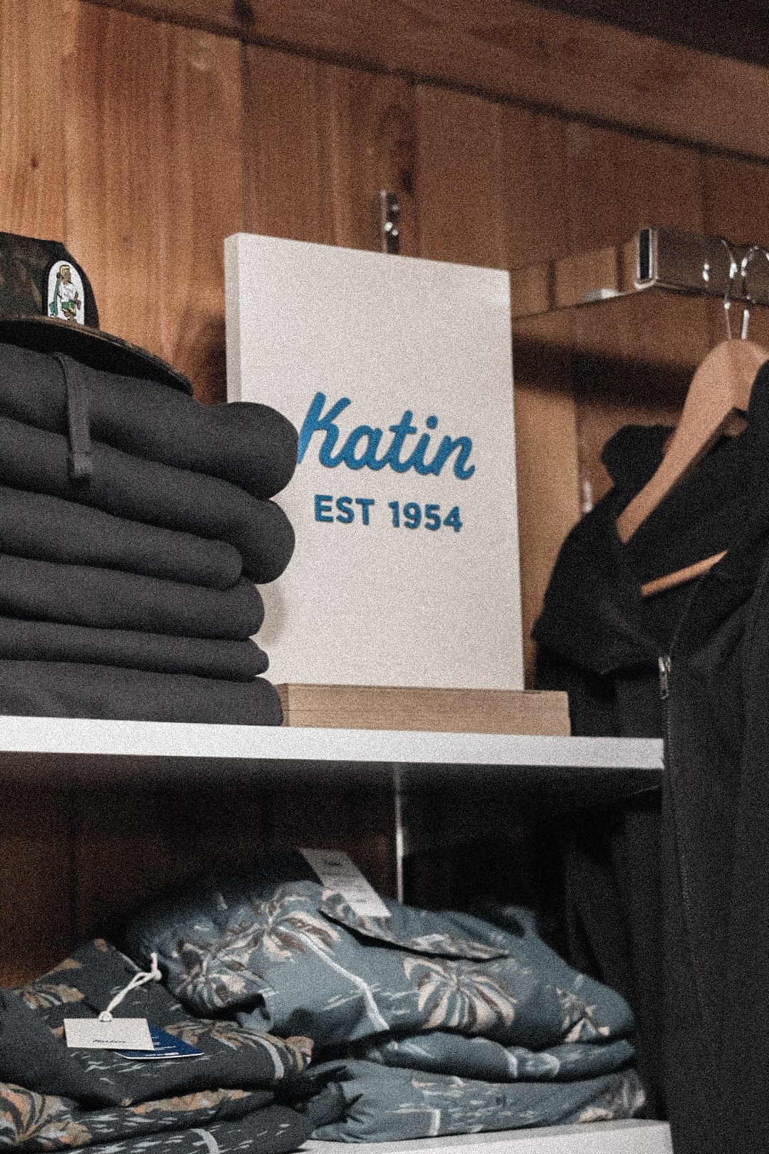 A retail display of black clothing, a sign with the text 'Katin EST 1954', and folding chairs with a floral pattern stored on a shelf.