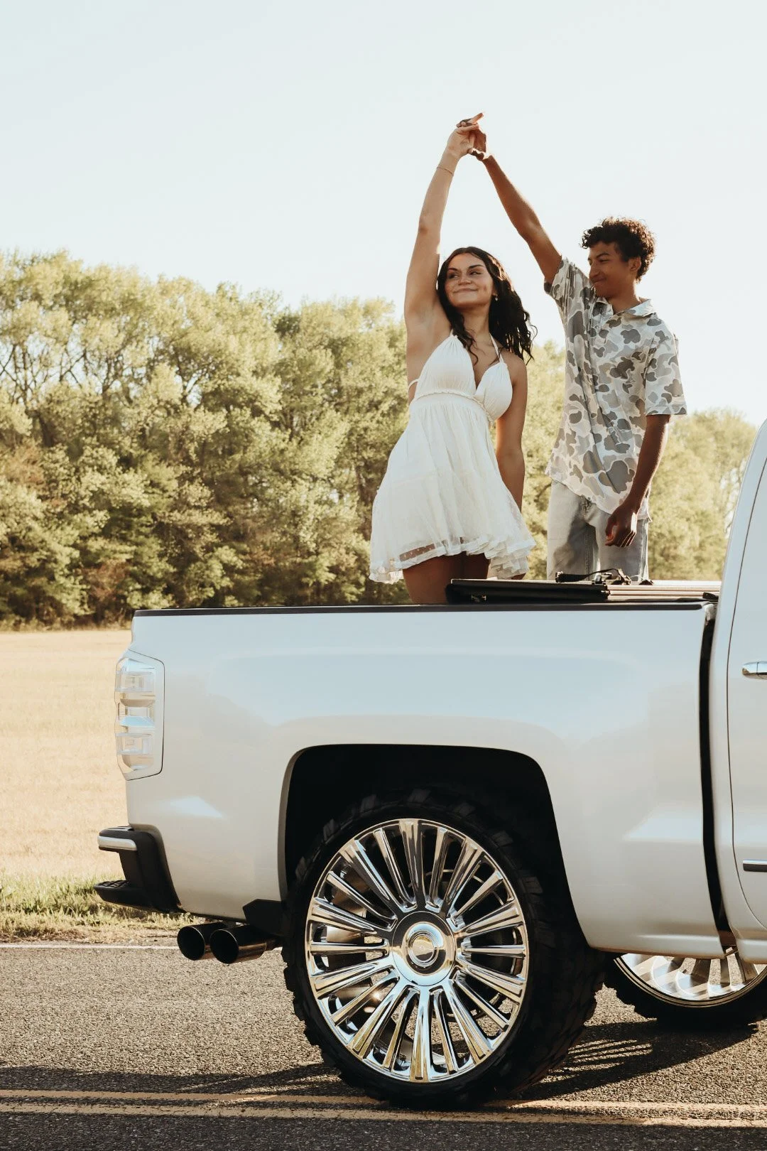 A woman and a boy dancing hand in hand in the back of a white pickup truck parked on a road, with trees in the background, during daylight.