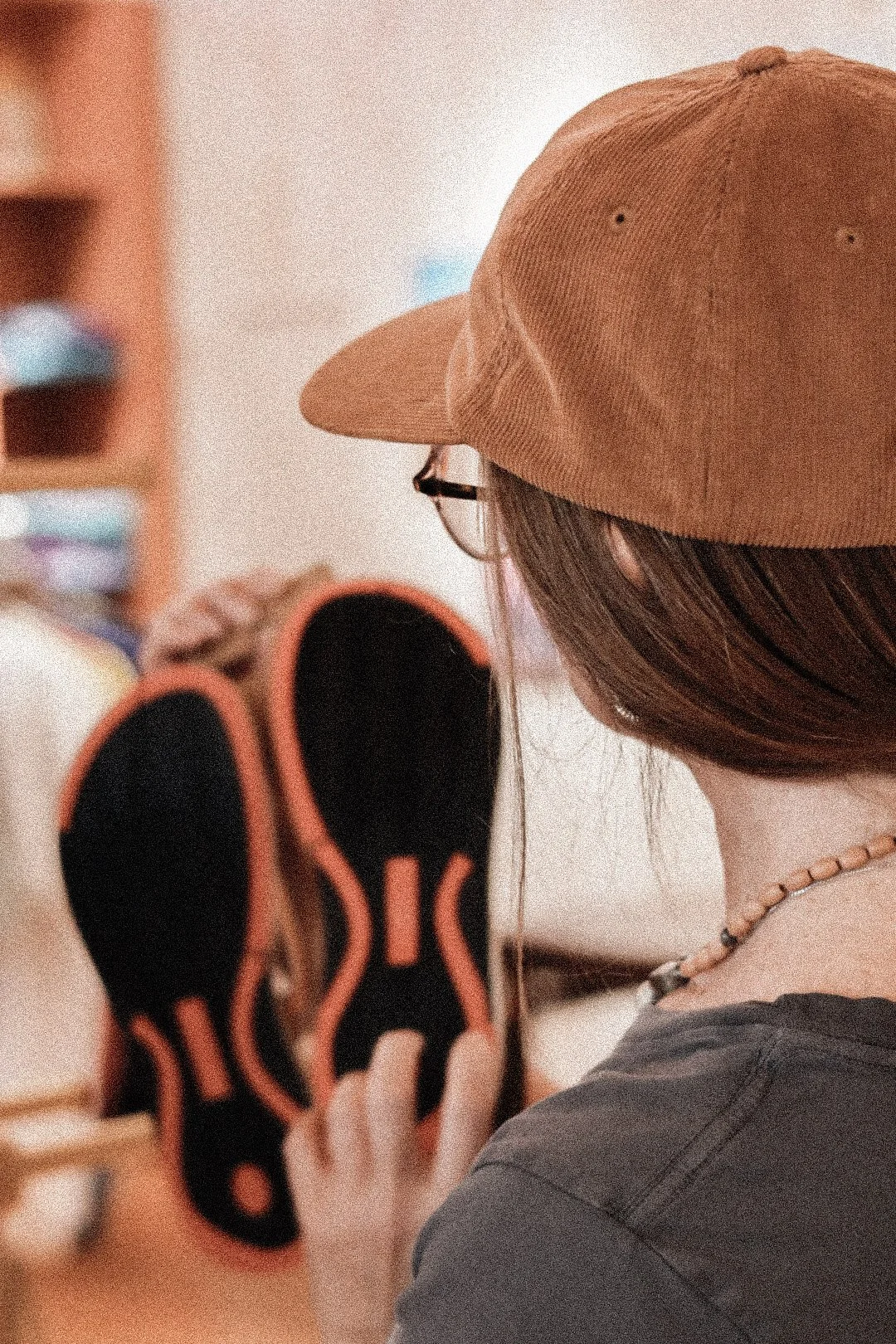 Person with long hair wearing glasses, a beige baseball cap, and a beaded necklace, holding a black and orange sneaker.