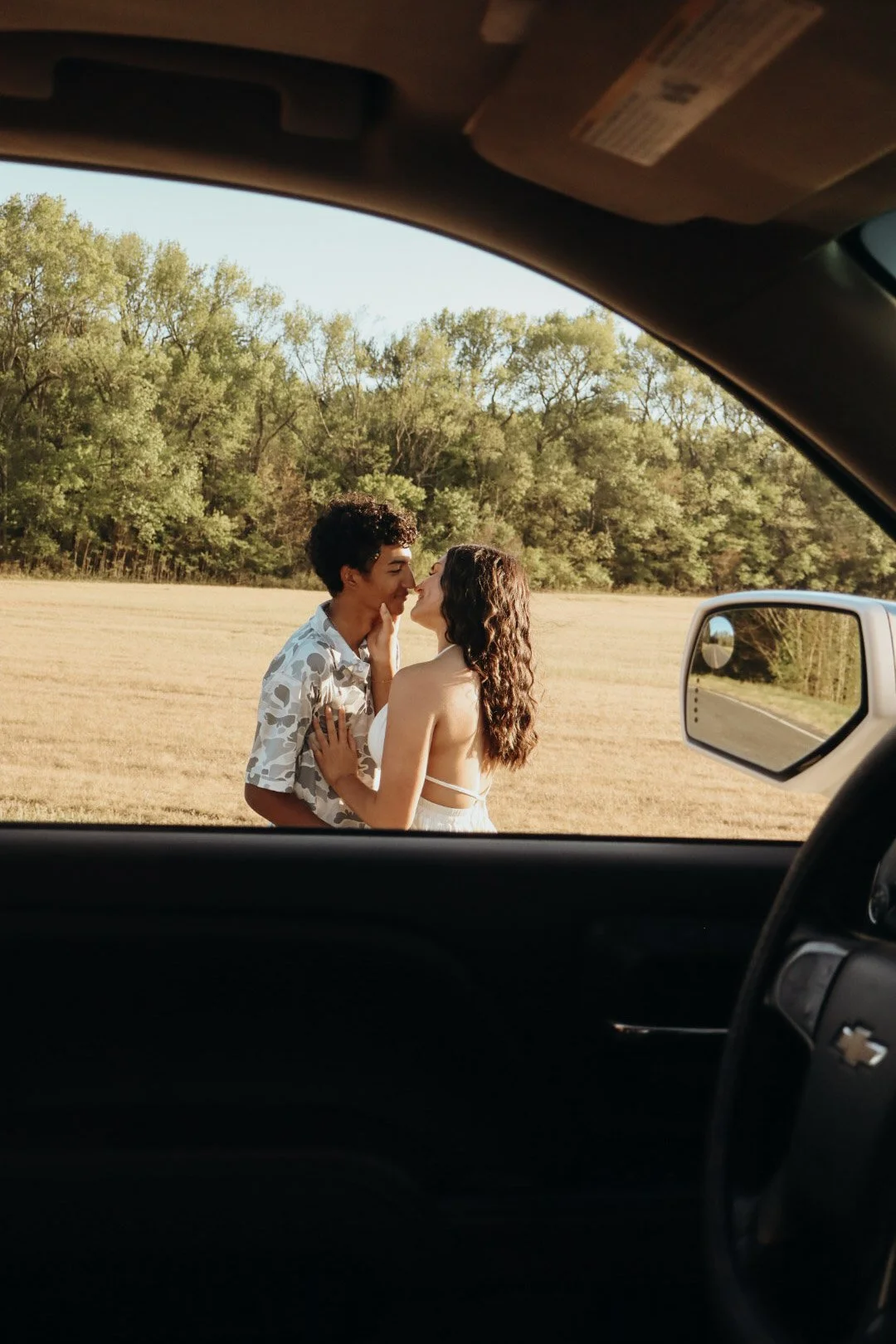 A couple is standing outside in a field, sharing a kiss, with trees in the background, seen through the open window of a car.